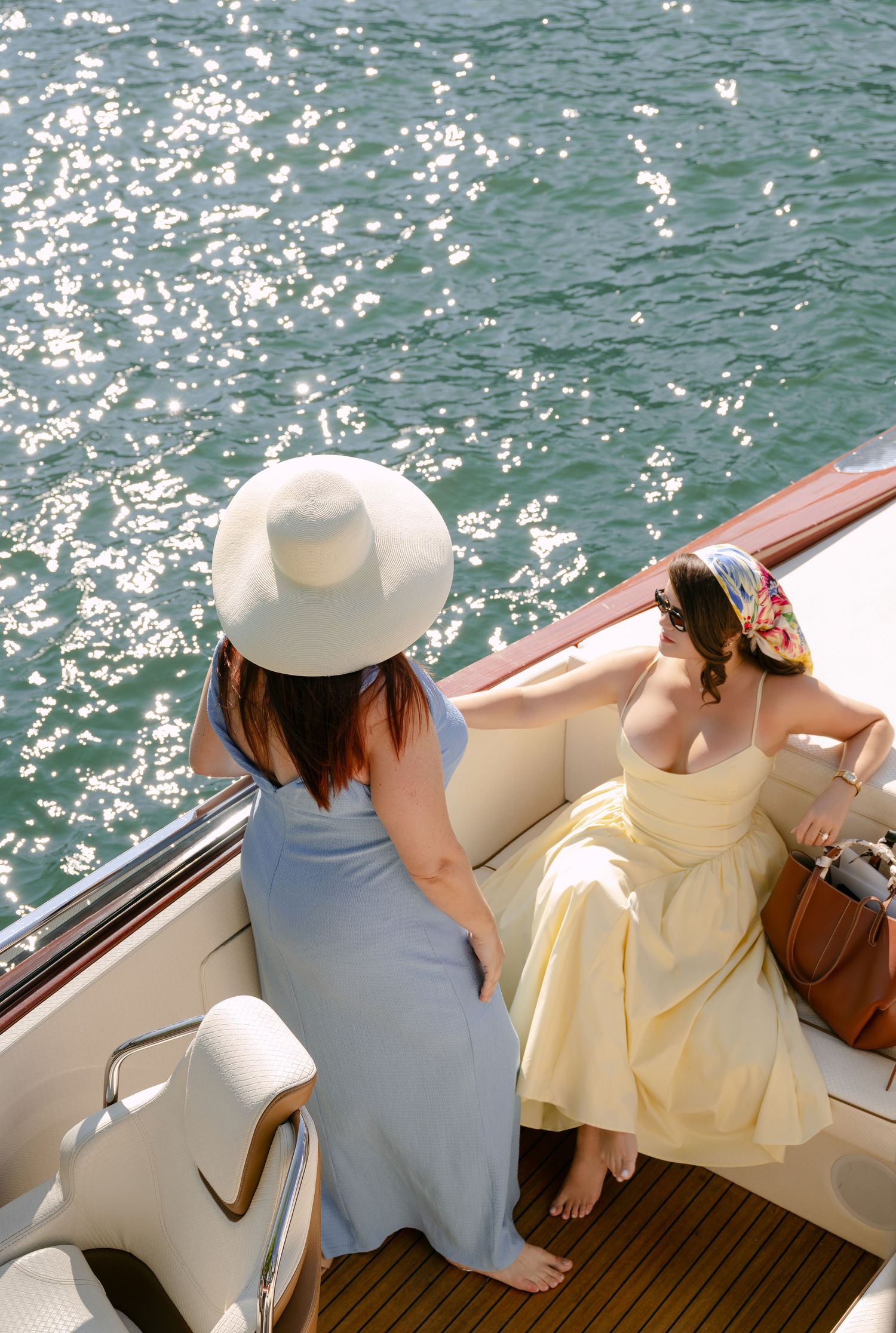 Two women in summer dresses relax on a luxury boat deck, one wearing a white sun hat, on sparkling water.