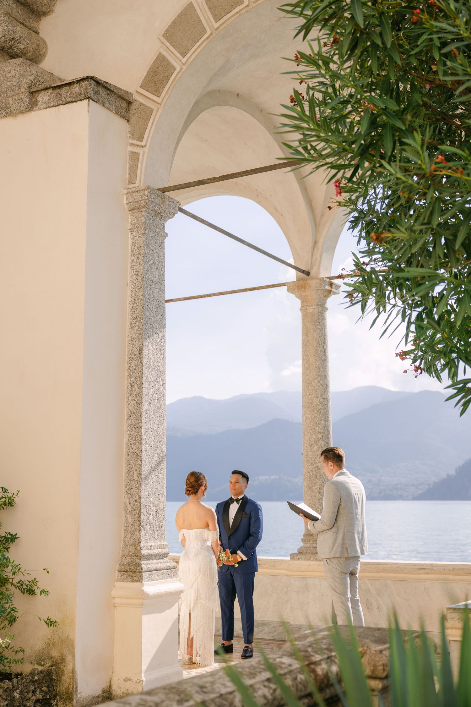 Couple exchanging vows at an elegant lakeside villa with arched columns and mountain views during an intimate outdoor wedding ceremony.
