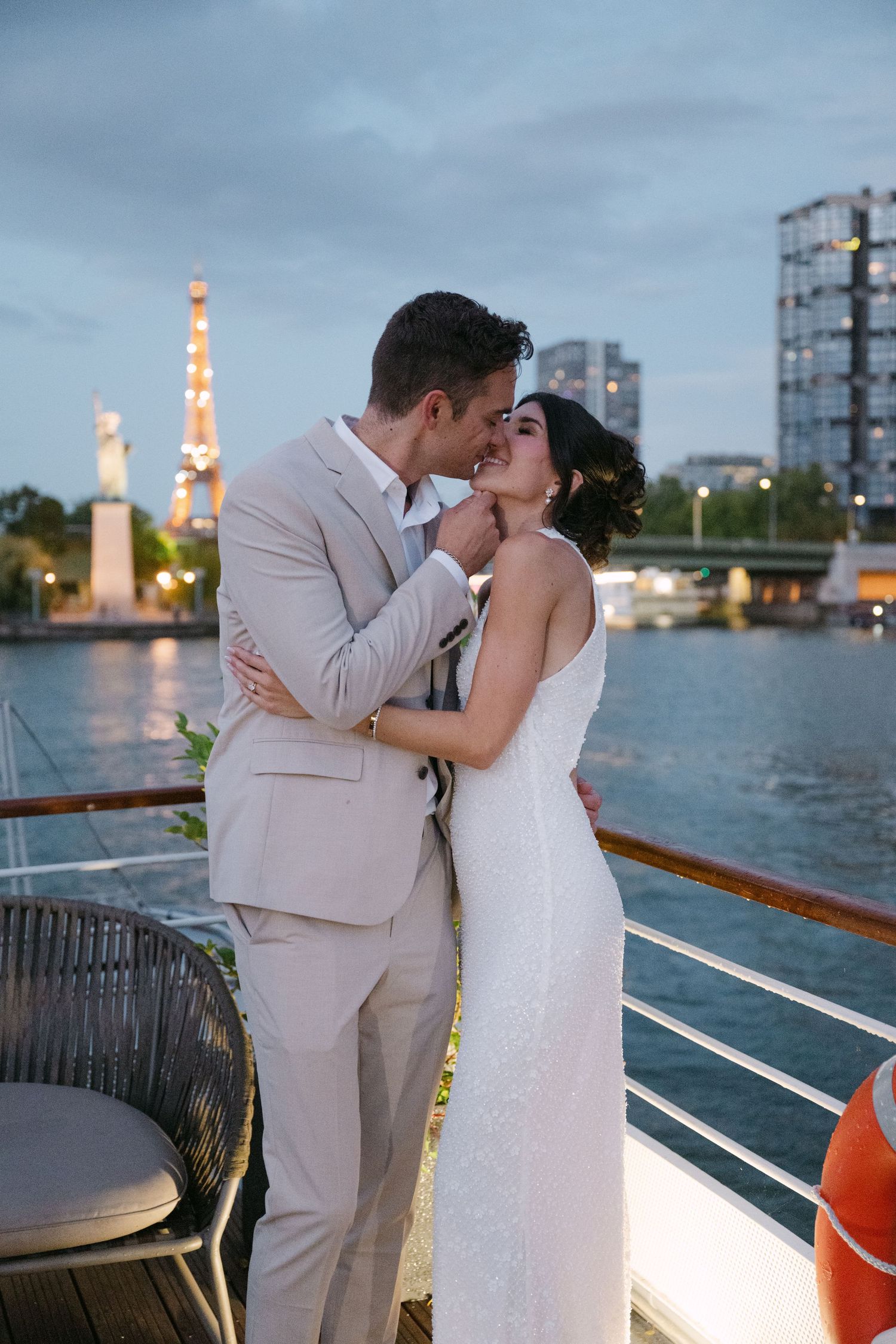 A bride and groom share a romantic kiss on a boat deck with the illuminated Eiffel Tower behind them.