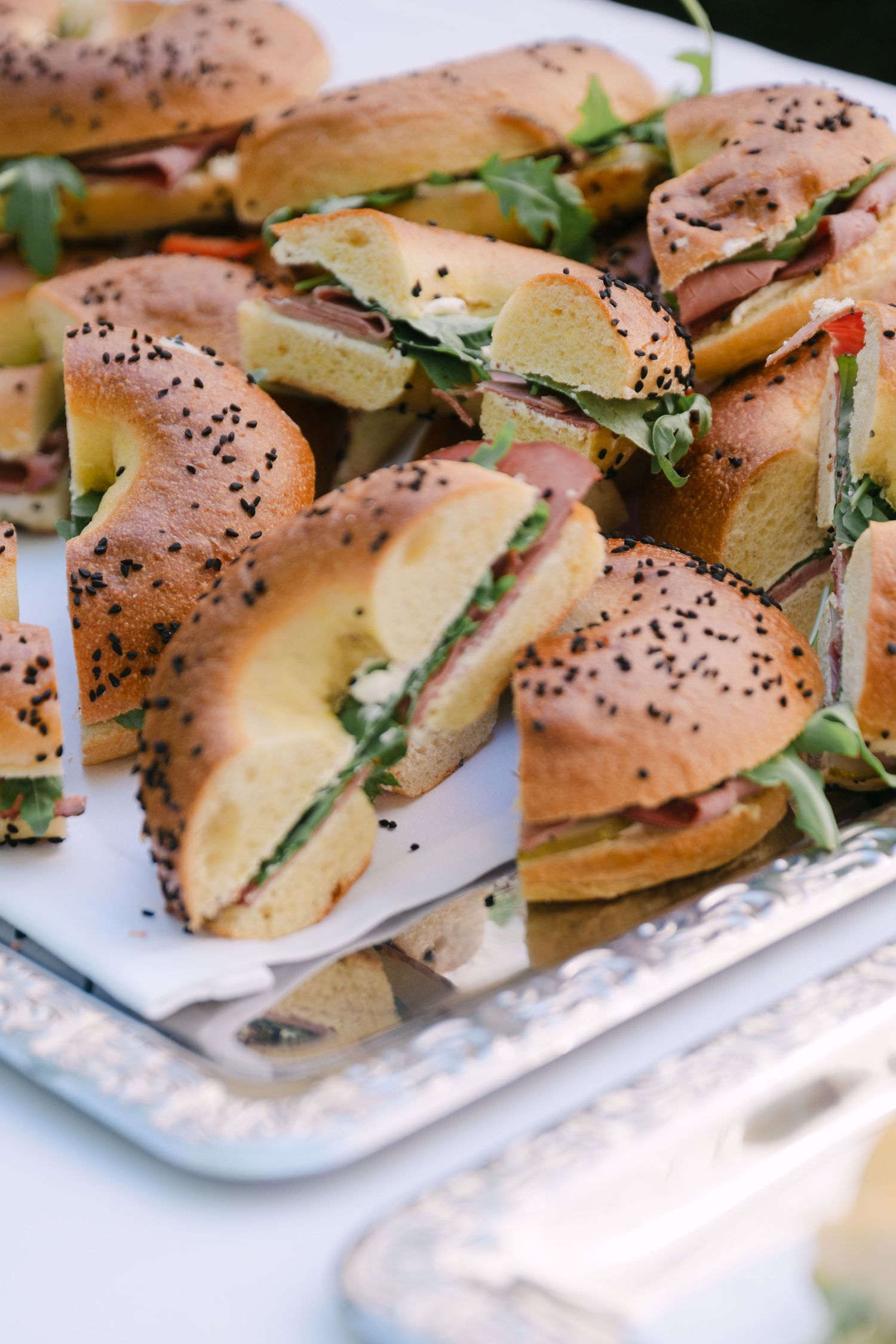 Tray of sesame seed bagel sandwiches filled with meat and arugula at a catered event.