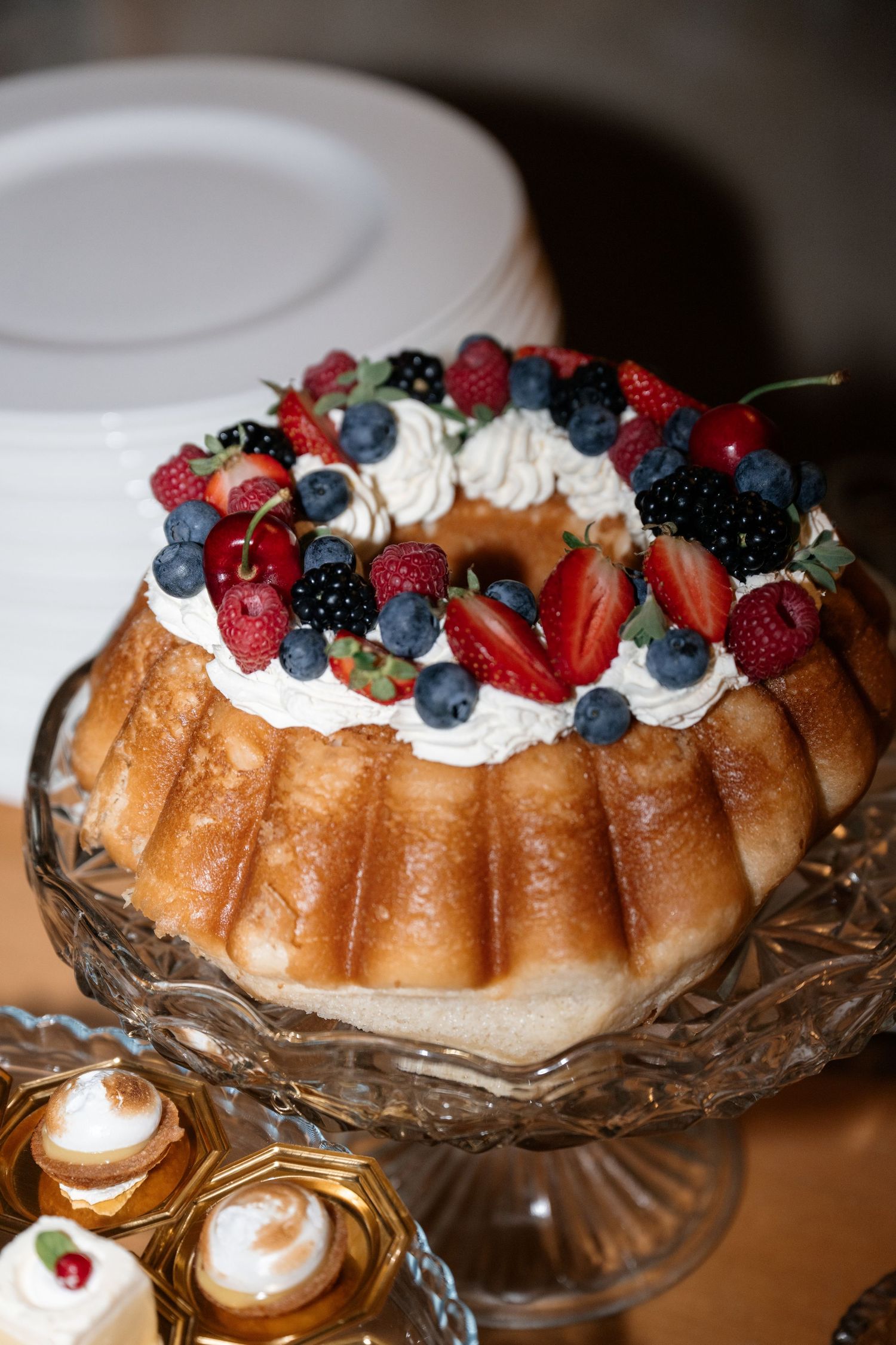 Elegant bundt cake topped with fresh berries and whipped cream on a crystal cake stand at a wedding dessert table.