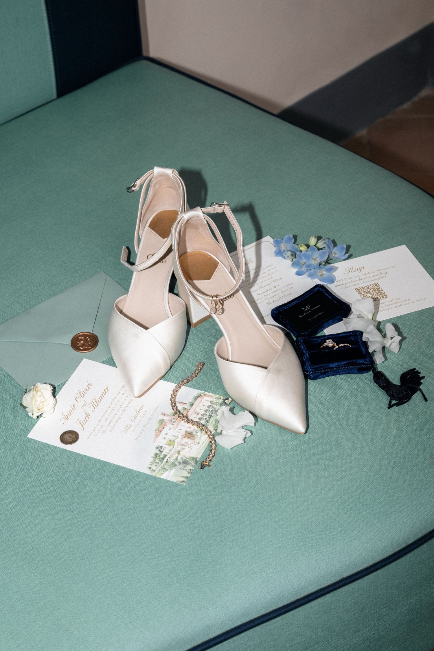 Elegant bridal flat lay with white heels, wedding invitation, ring box, and floral accessories on teal surface.