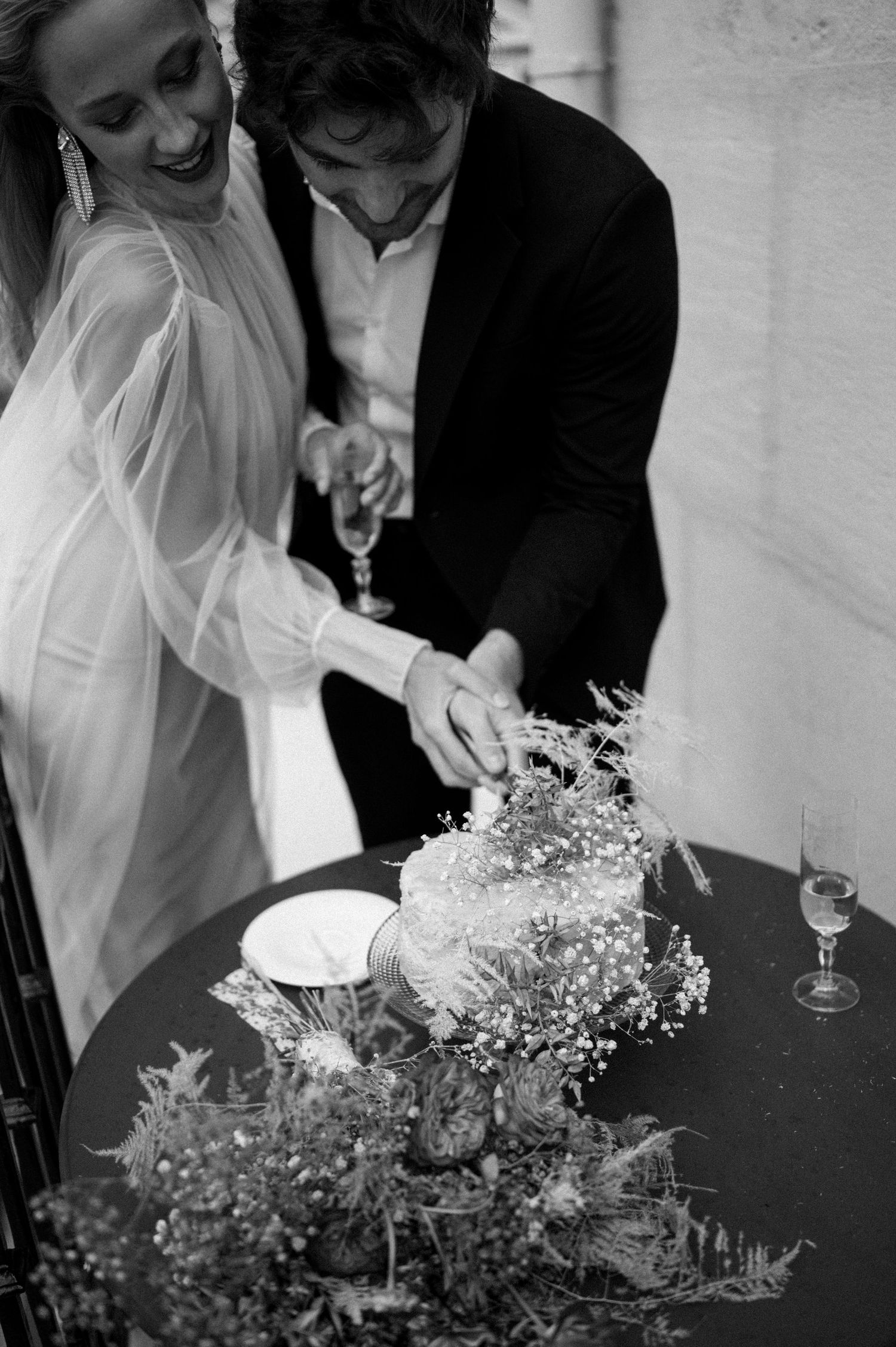 Black and white photo of smiling newlyweds cutting their flower-decorated wedding cake together on a balcony.
