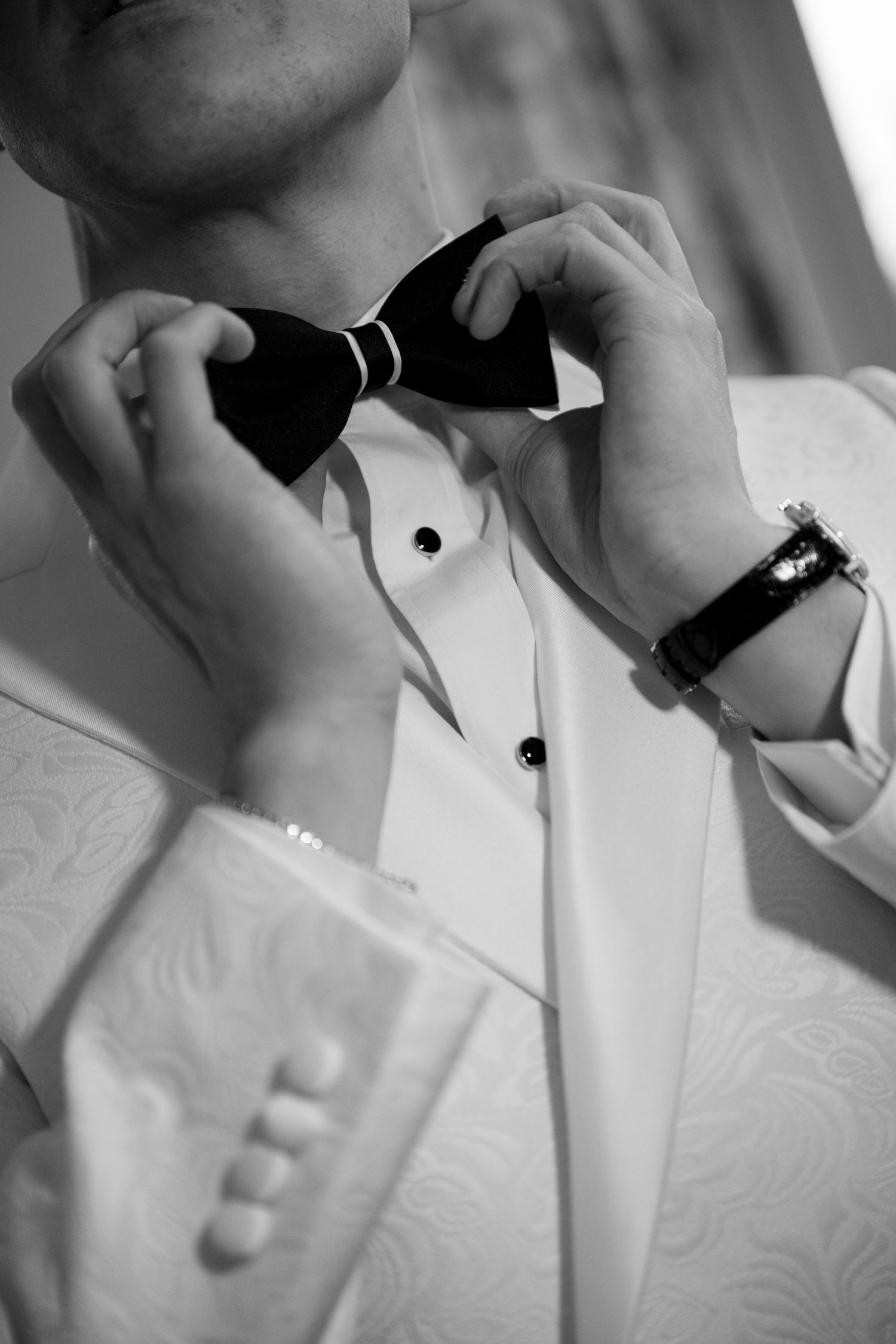 Black and white close-up of groom adjusting black bow tie with watch on wrist.