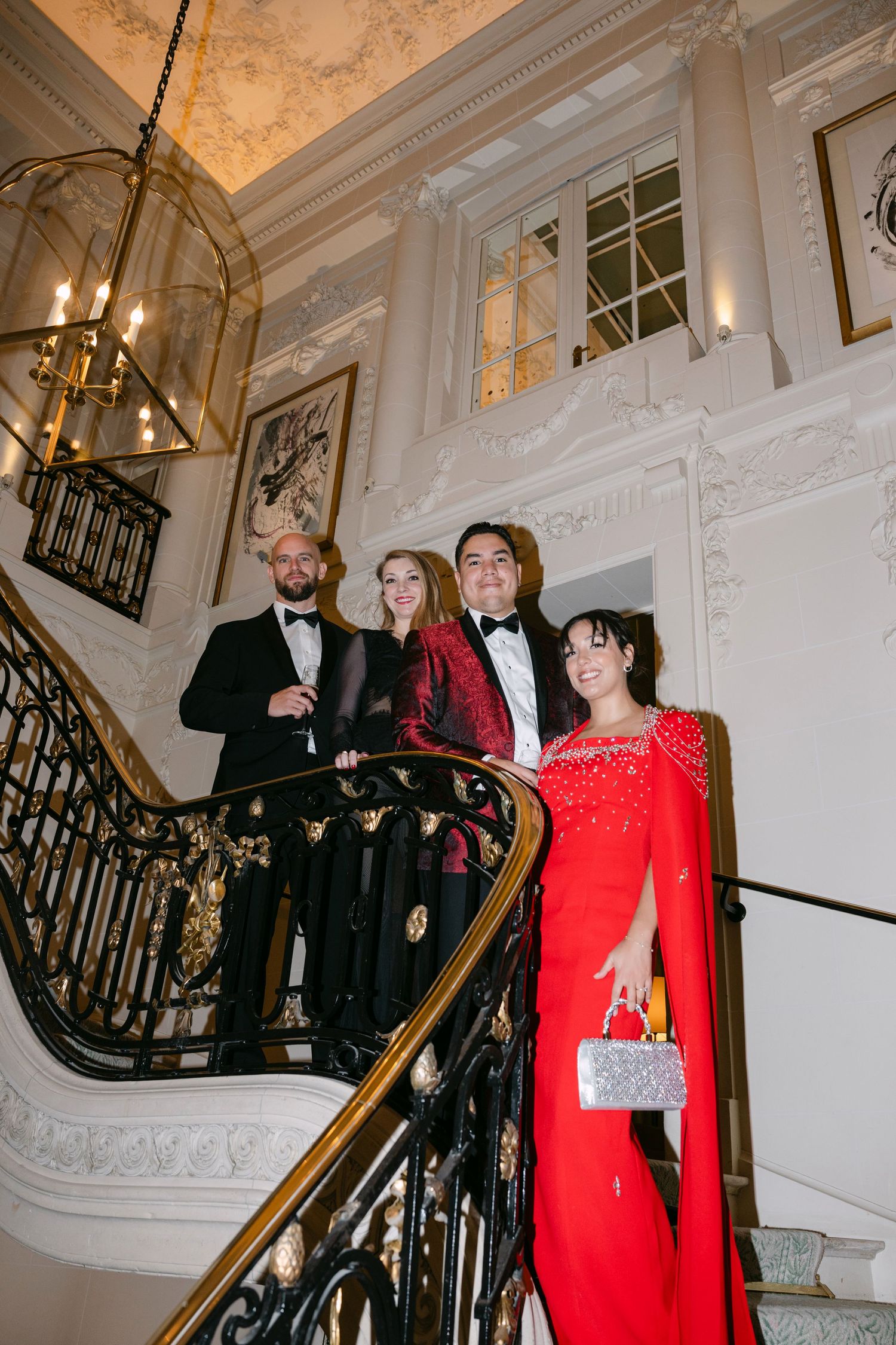 Four elegantly dressed wedding guests pose on ornate grand staircase in opulent venue, two women in red and burgundy gowns.