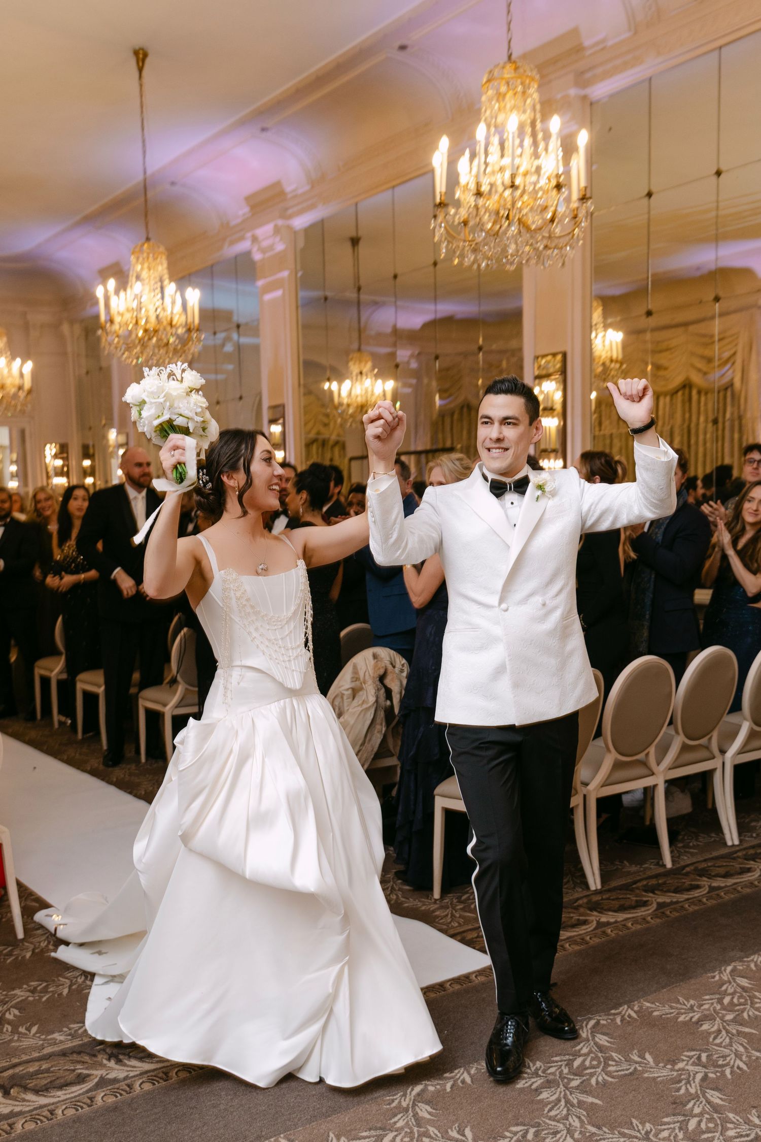 Joyful newlyweds celebrate grand entrance at luxurious ballroom wedding, bride in white gown raising bouquet with groom in white tuxedo.