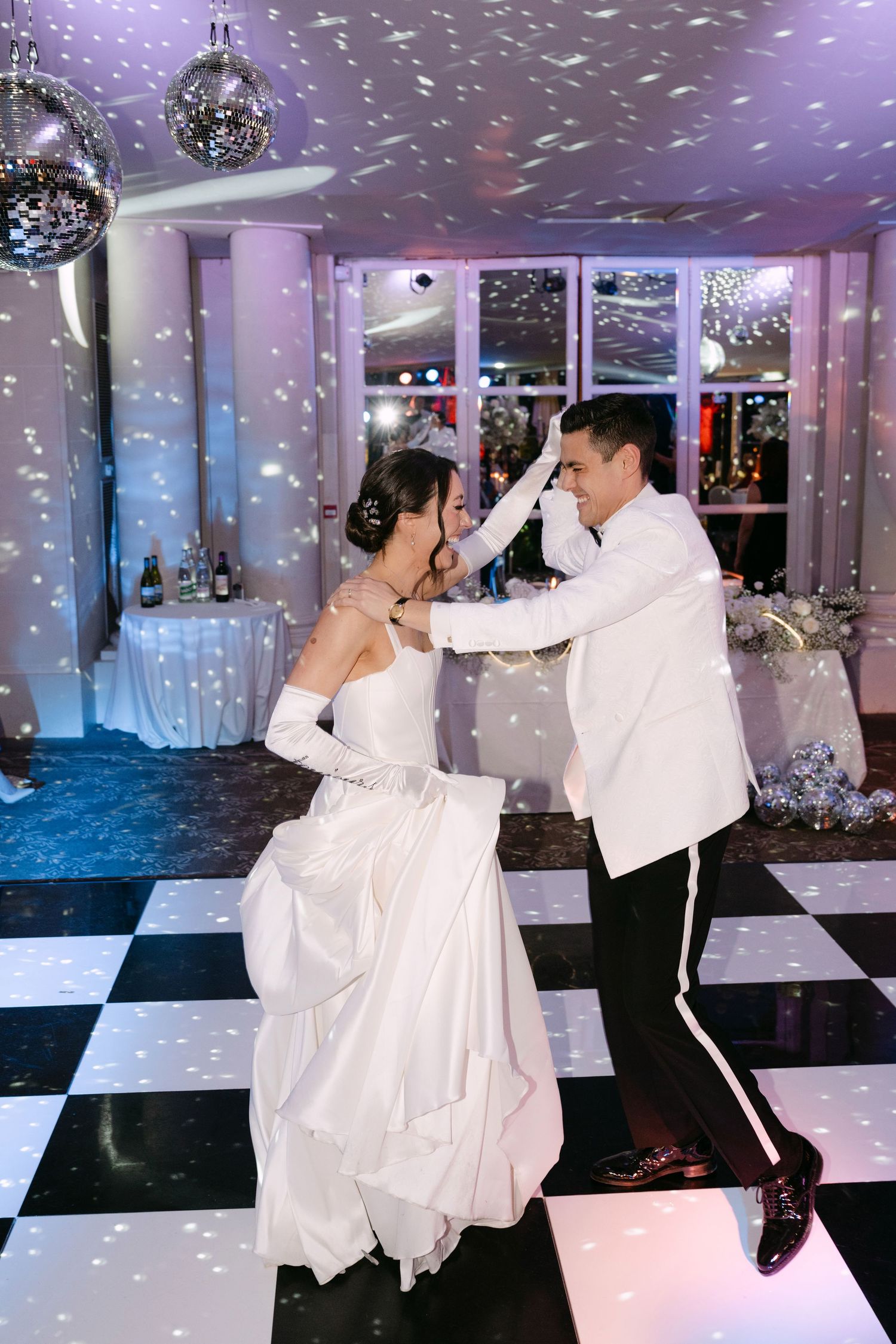 Bride and groom share their first dance on a black and white checkered floor under sparkling disco balls.
