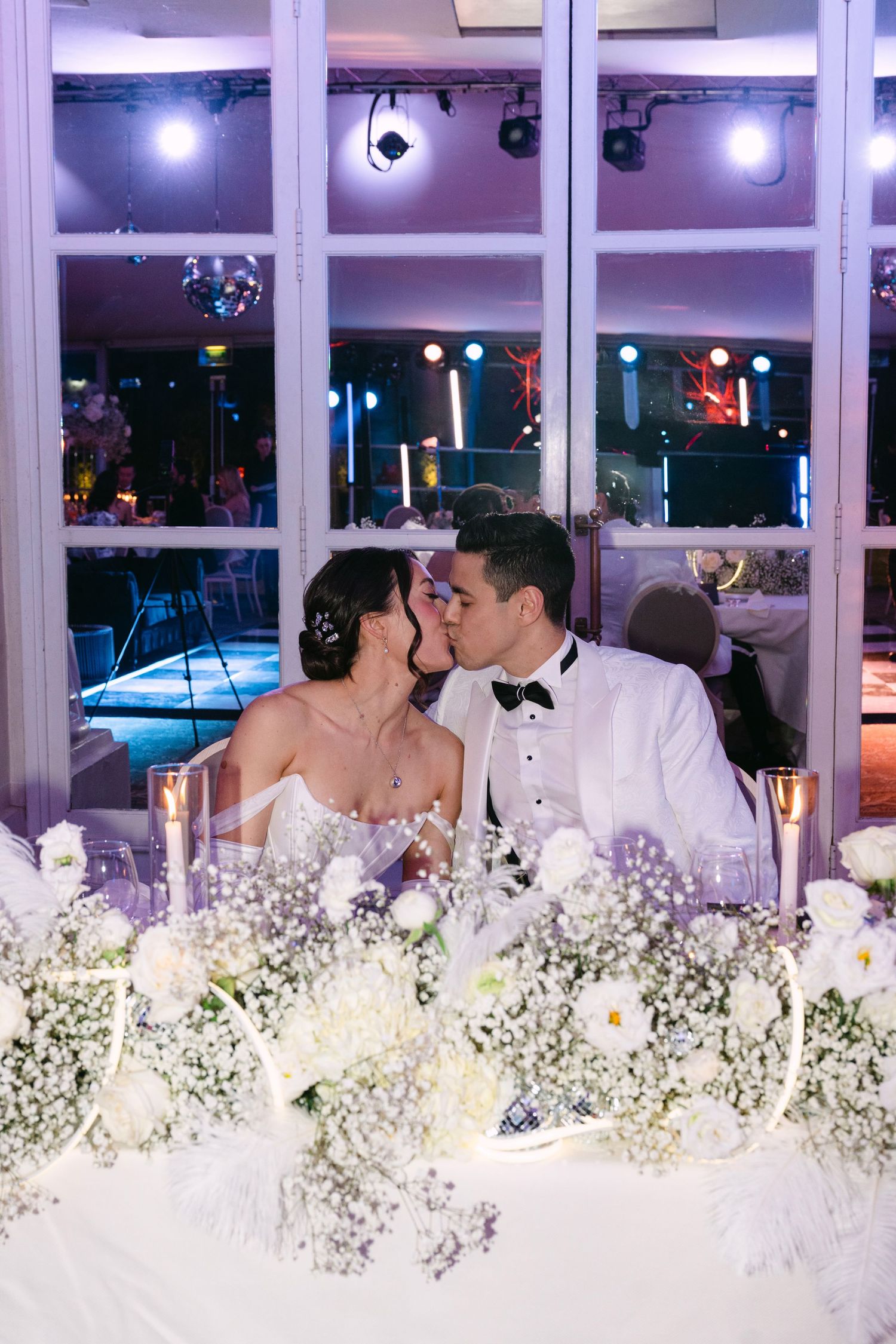 Newlyweds share romantic kiss at sweetheart table adorned with white roses and baby's breath.