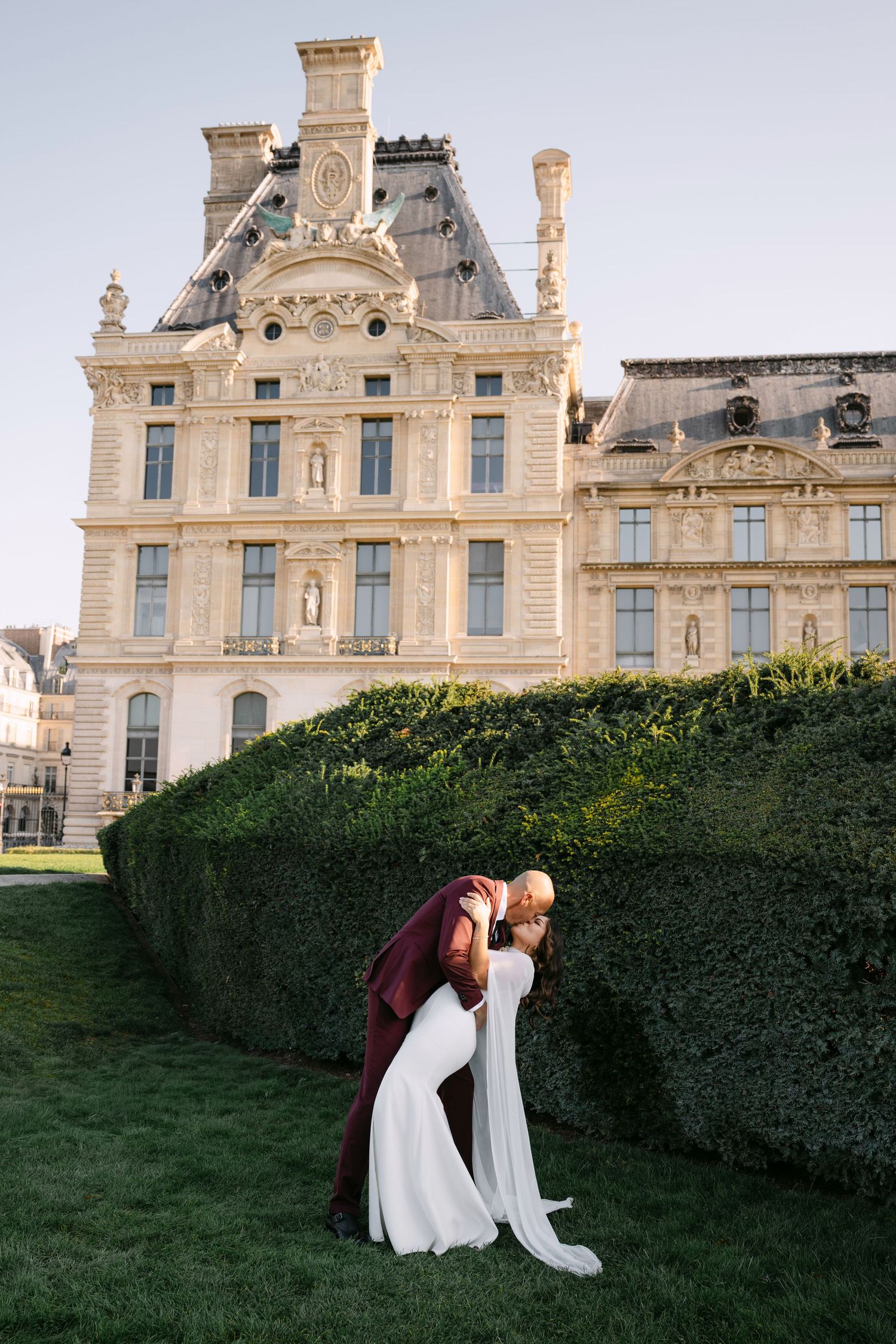 Couple shares romantic dip kiss beside manicured hedge with elegant Louvre building in background.