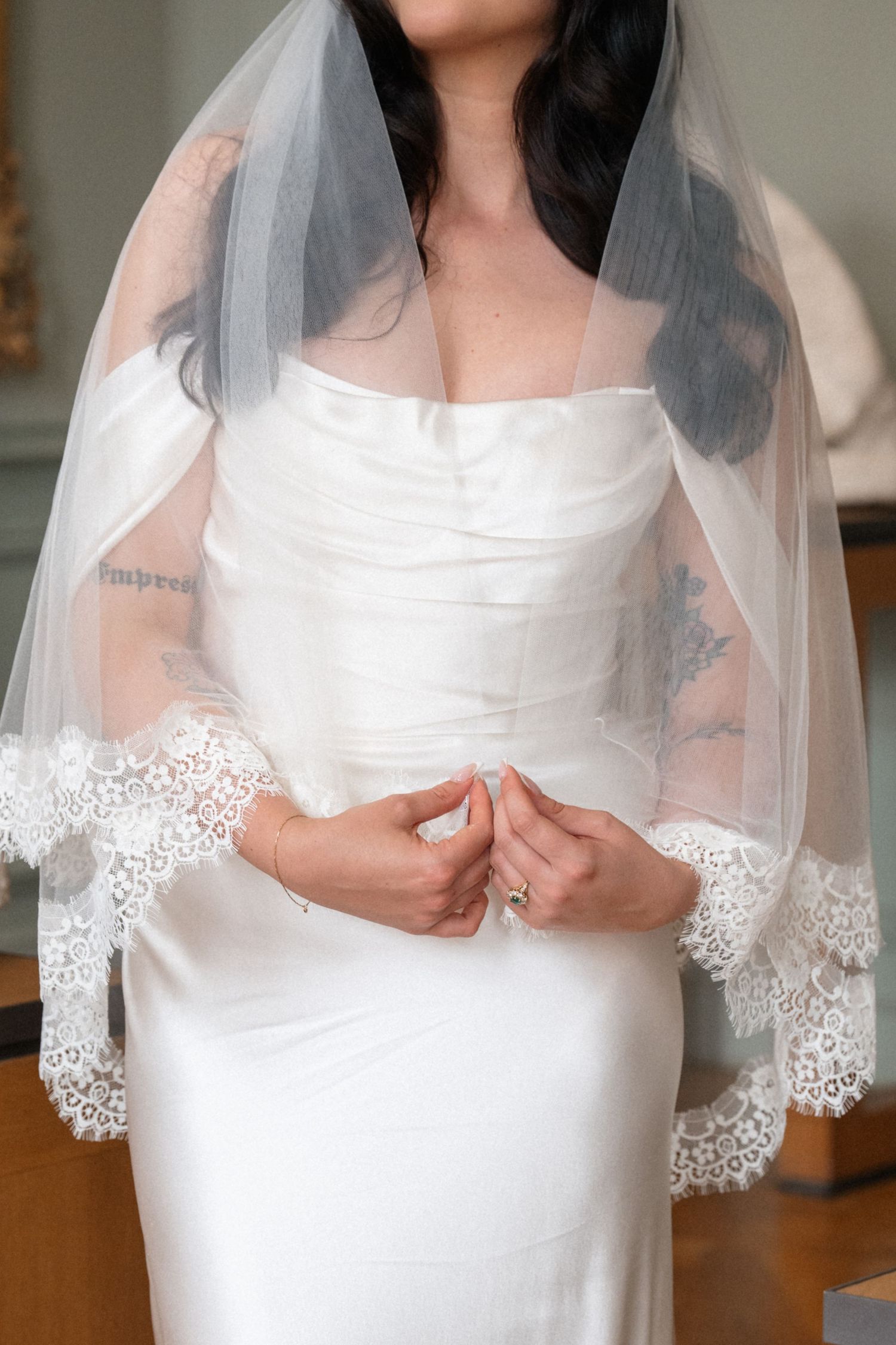Close-up of bride in off-shoulder satin wedding gown with lace-trimmed veil and visible arm tattoos.