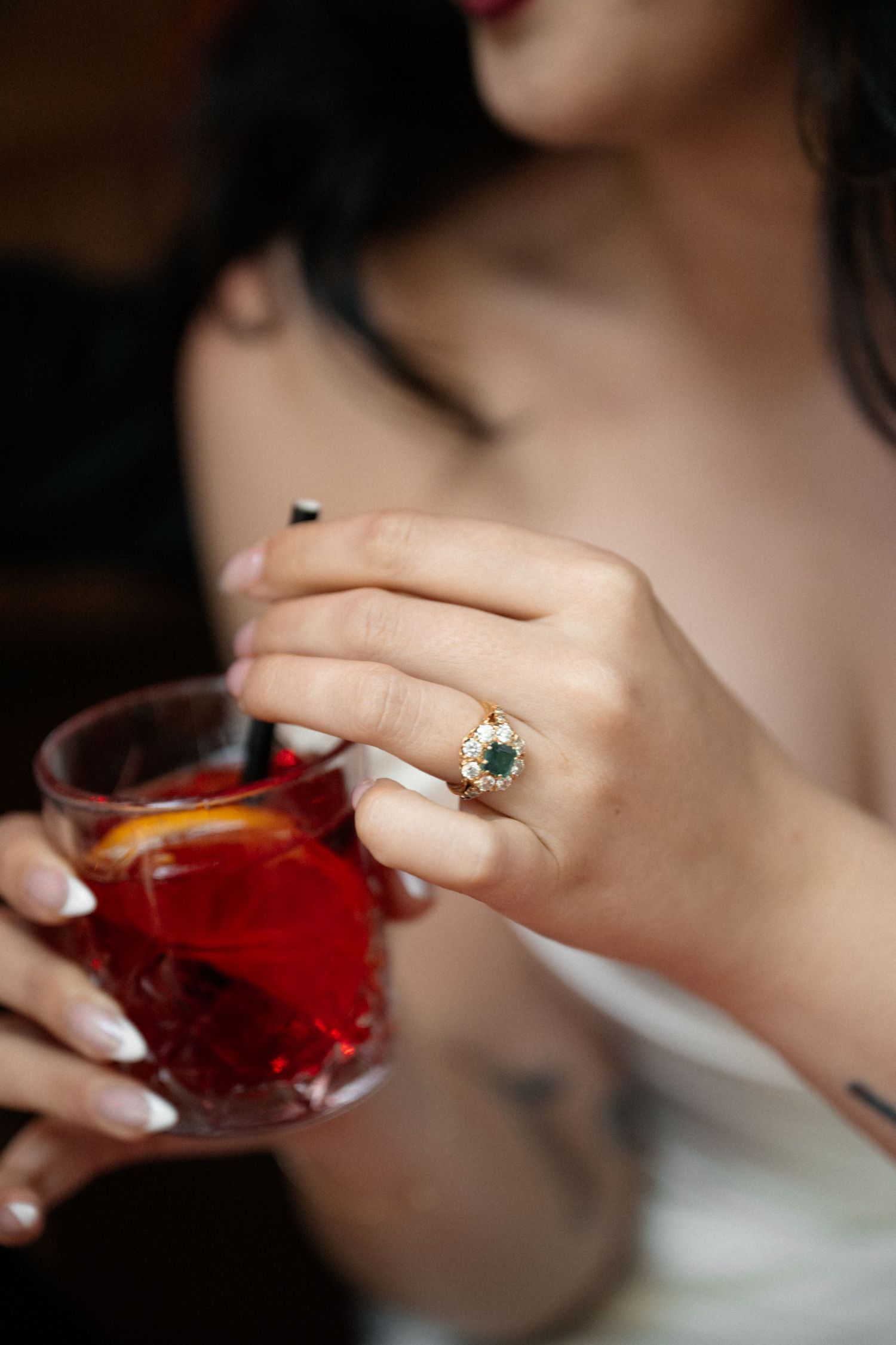 Close-up of bride wearing emerald and diamond halo engagement ring while holding a Negroni cocktail.