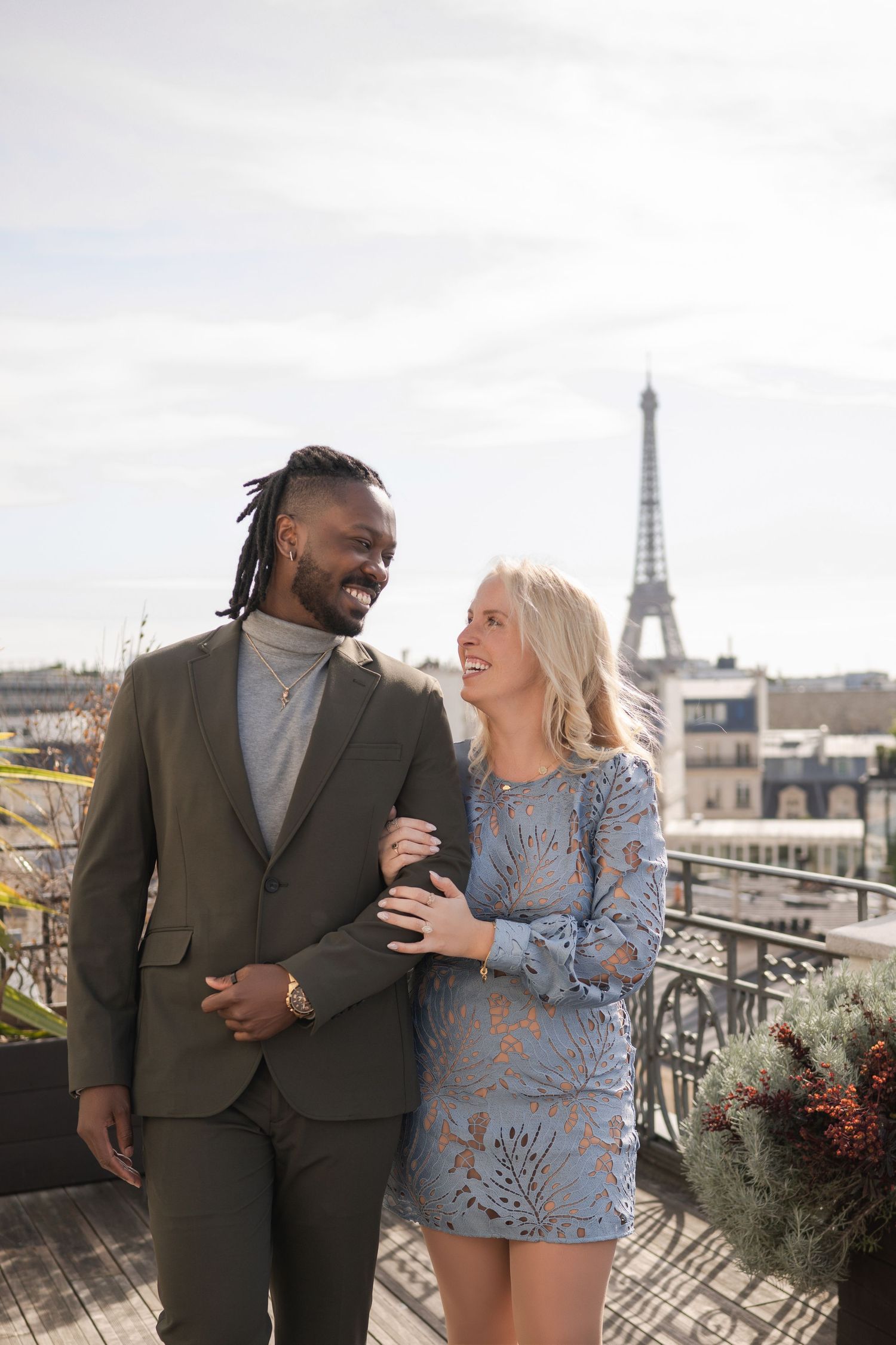 A smiling couple poses on a Paris rooftop terrace with the Eiffel Tower visible in the background.