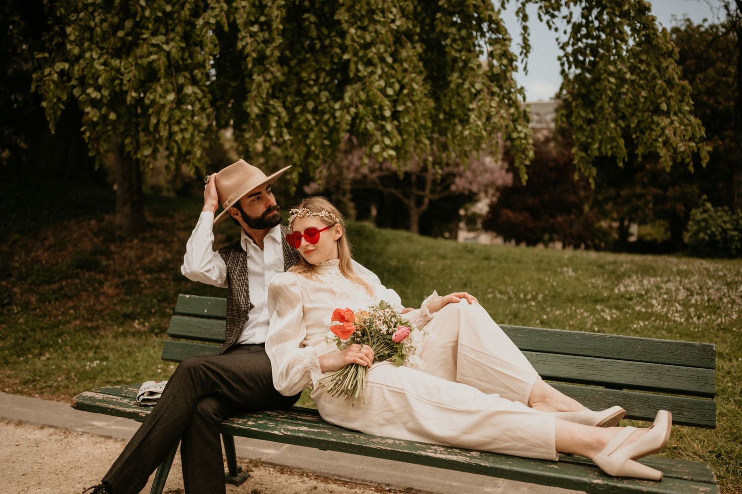 A stylish pair lounges on a green park bench surrounded by lush trees in retro-inspired outfits.