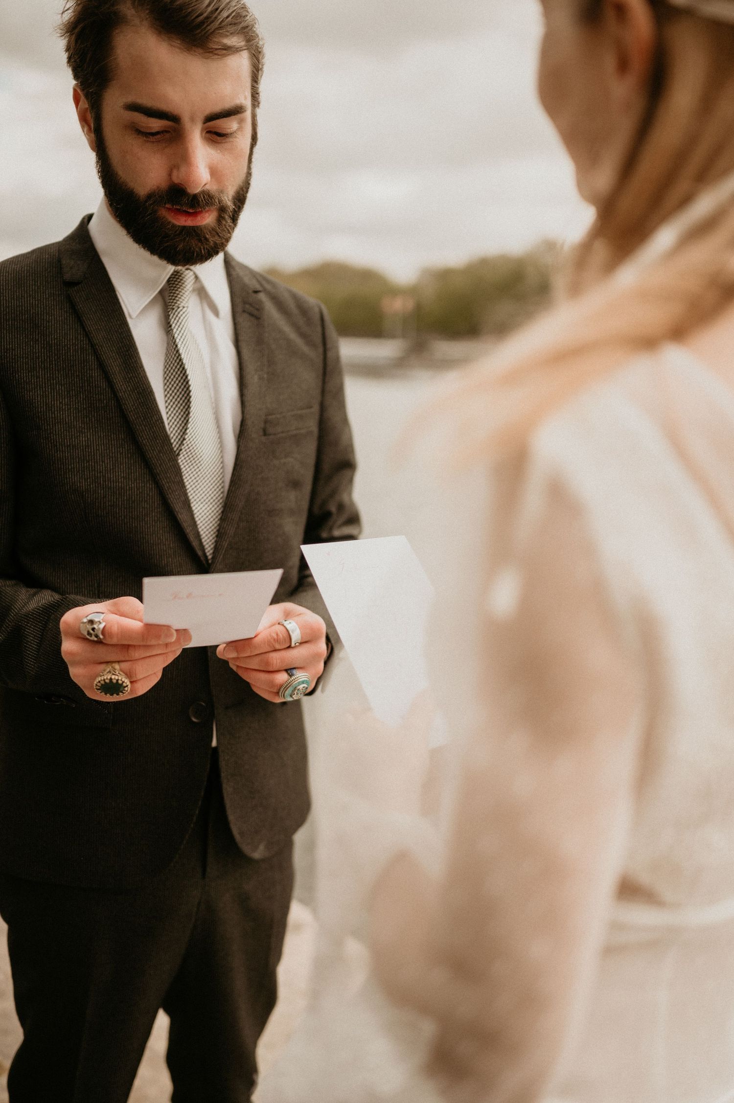 A groom in a dark suit reads personal vows during an outdoor wedding ceremony.