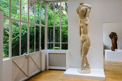 Modern gallery space with large windows displaying classical nude sculpture and natural light.