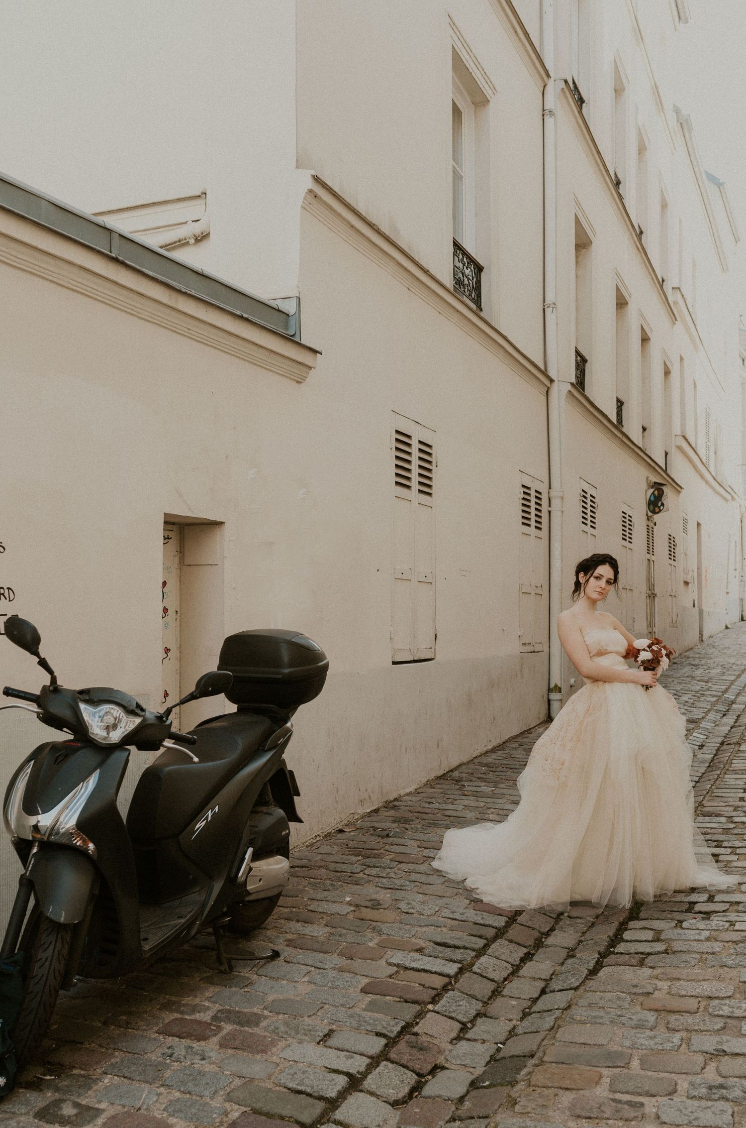 Bride in tulle gown stands near parked scooter on narrow cobblestone alley in Paris.