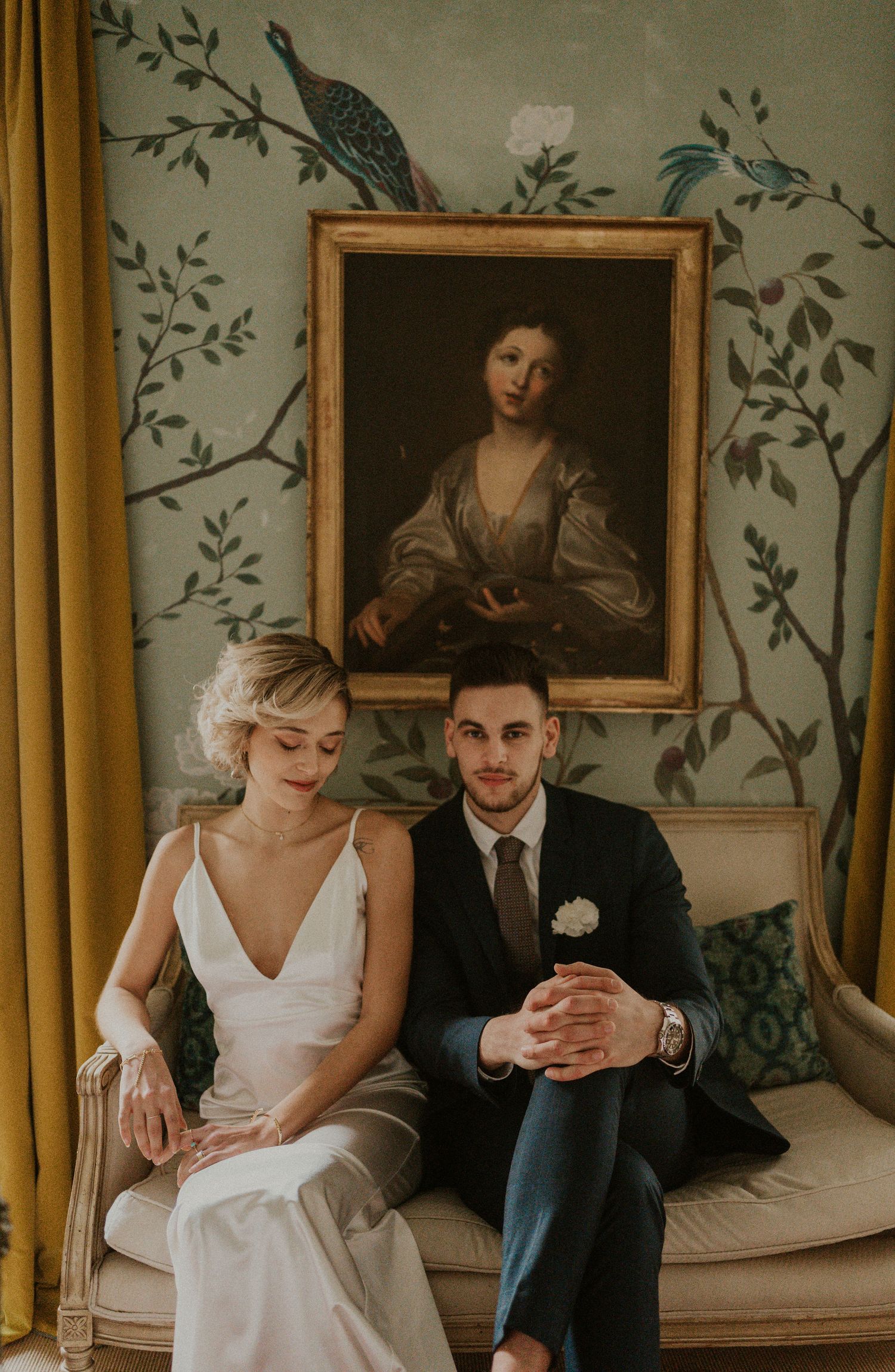 Wedding couple poses beneath vintage portrait in room with bird-patterned wallpaper.