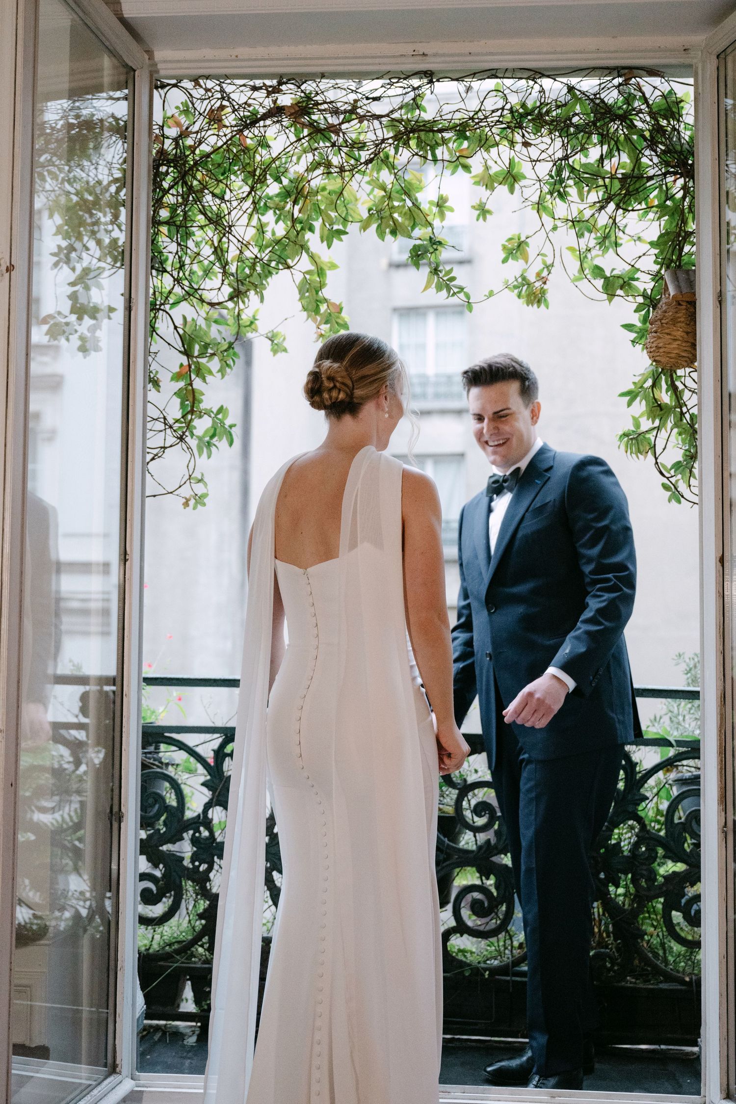 First look wedding moment captured on an elegant covered balcony with greenery and decorative railings.