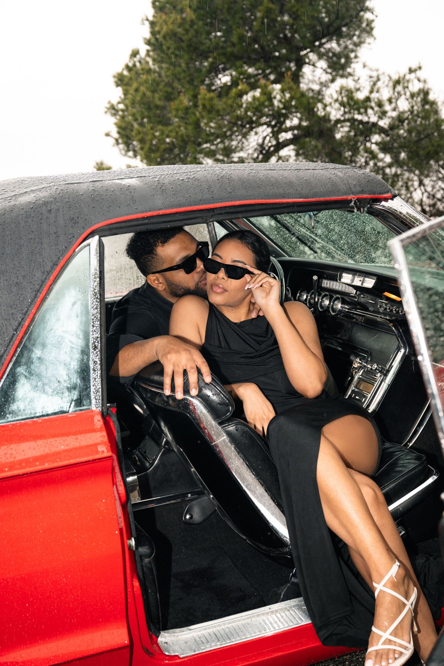 A couple in formal attire poses on a road next to a vintage red classic car with trees in the background.