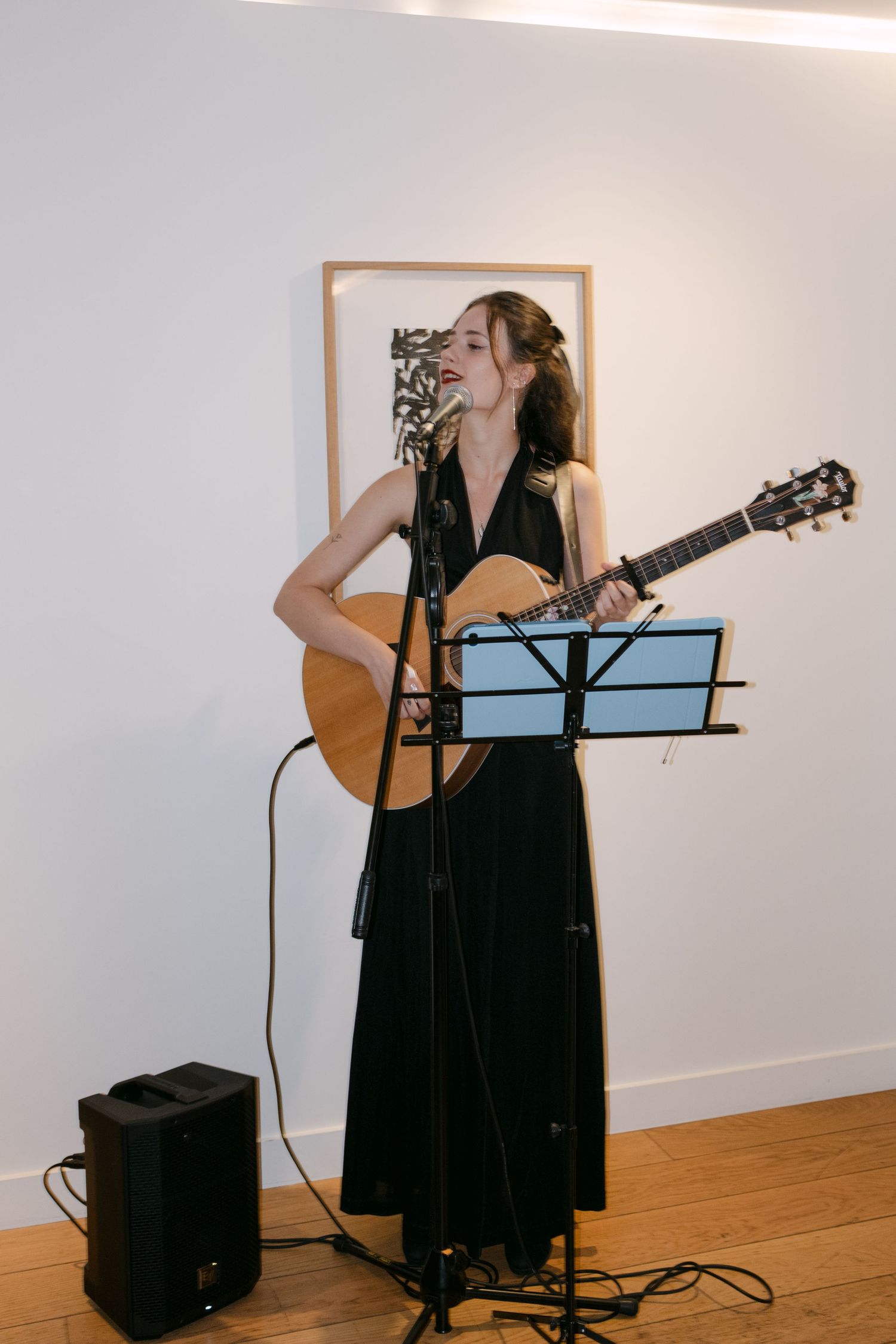 A musician in a black dress performs with an acoustic guitar and microphone stand against a white wall.