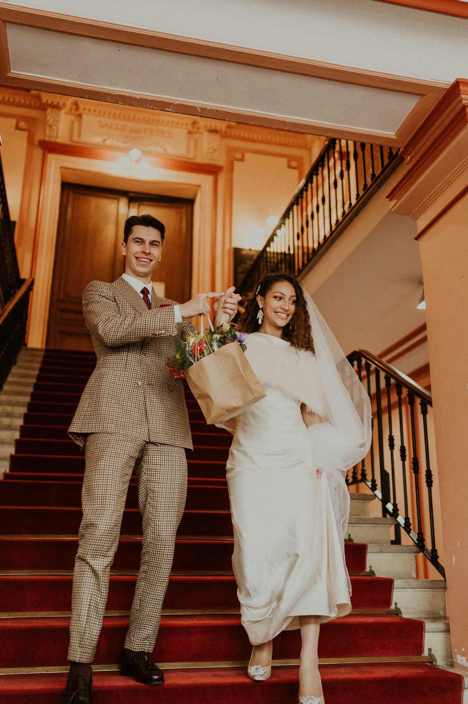 A happy couple descends grand stairs carrying flowers and smiling in formal attire.