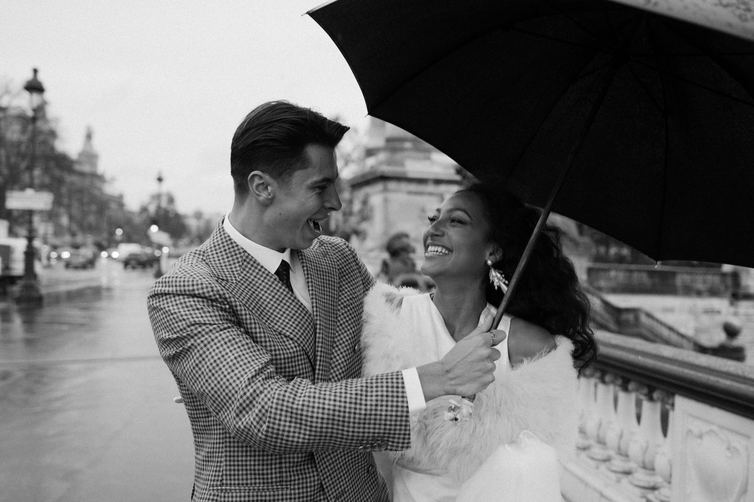 Black and white photo of couple sharing romantic moment under umbrella by ornate railing.