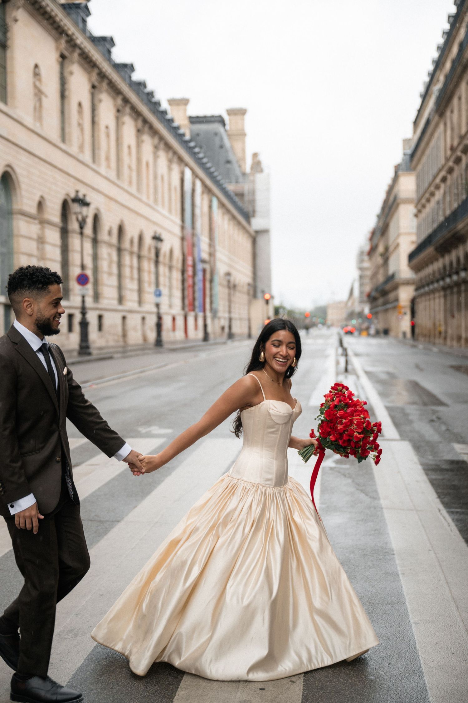 Bride in champagne gown holds red bouquet while walking hand-in-hand with groom on a Parisian street.