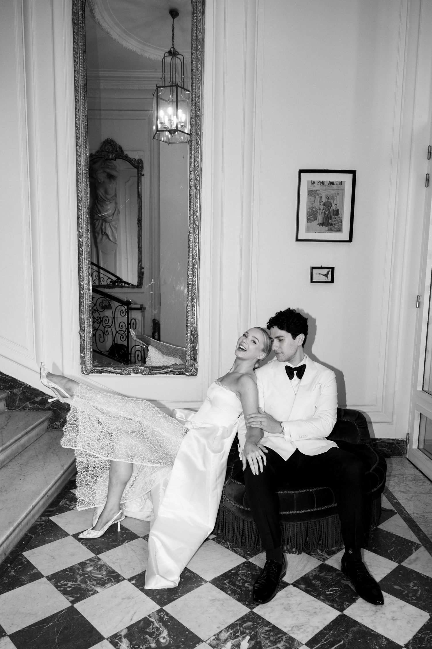 Black and white photo of bride and groom posing playfully on a velvet chair beside an ornate gilded mirror.