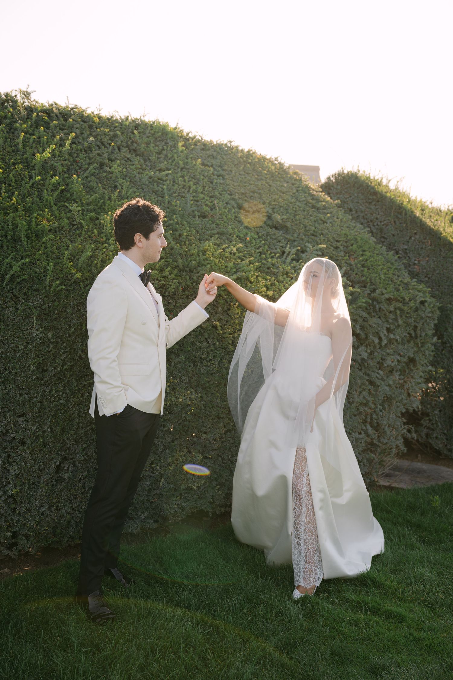 Groom in white jacket holds brides hand as she twirls in lace wedding gown and veil before a manicured green hedge.