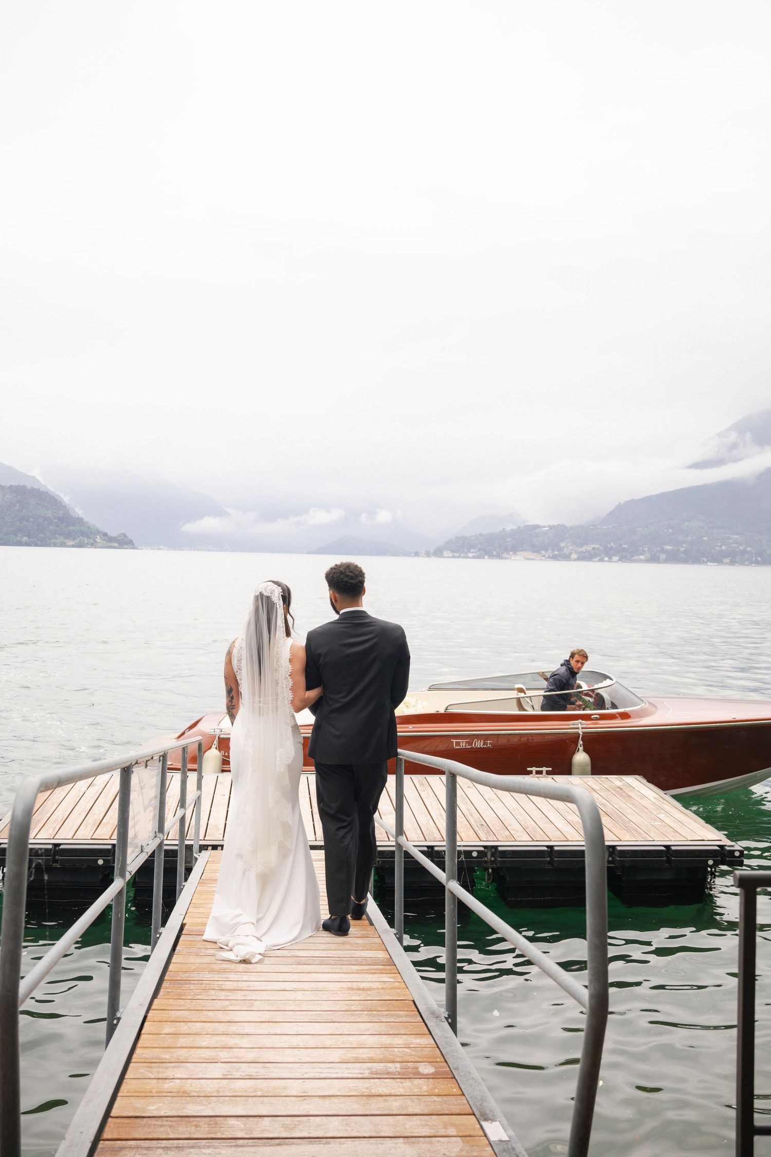 Bride and groom standing on a wooden dock overlooking a misty mountain lake with a classic wooden boat.