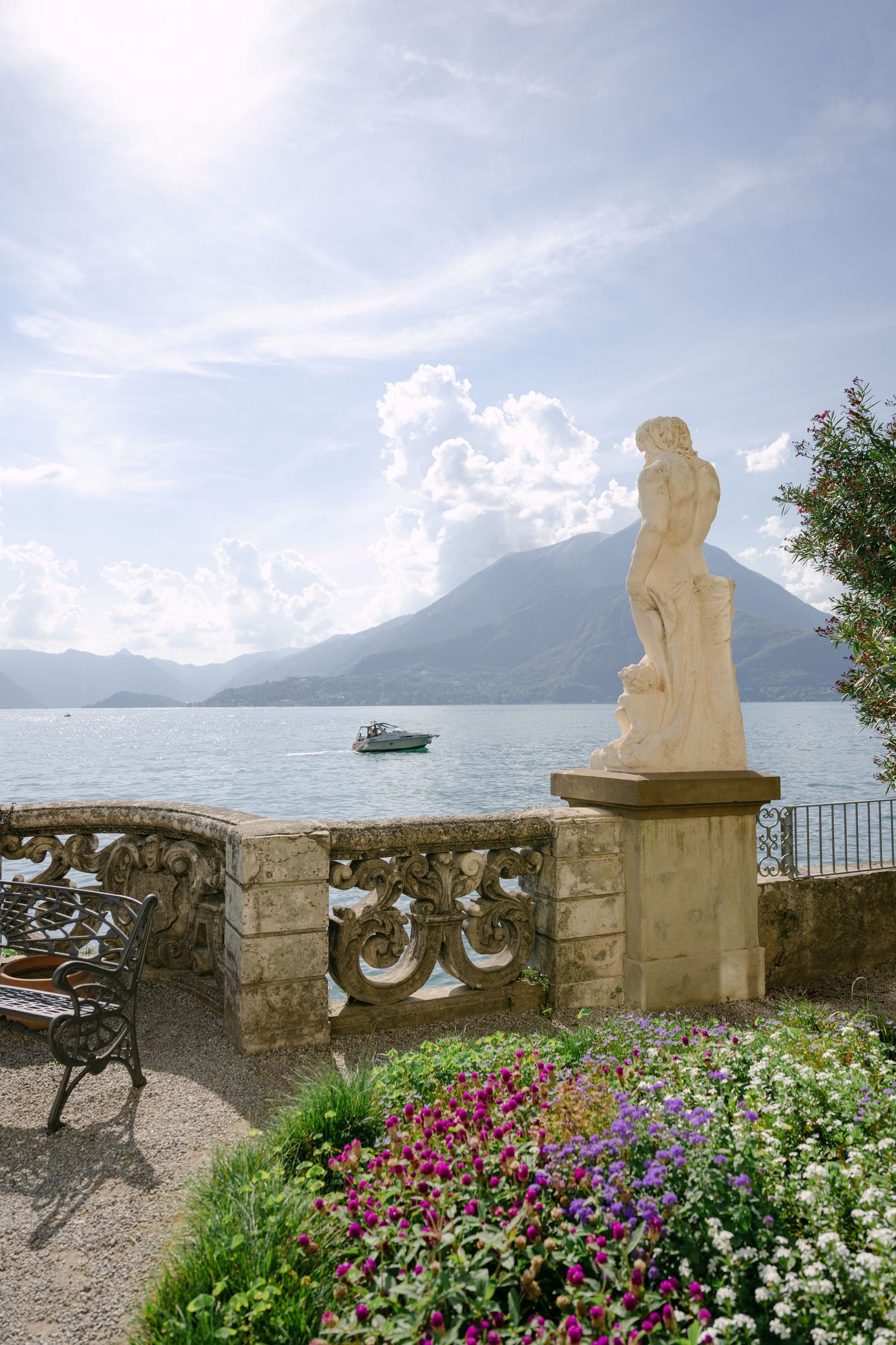 Classic marble statue on a terrace overlooking Lake Como, with colorful flowers, mountains, and a boat on the water.