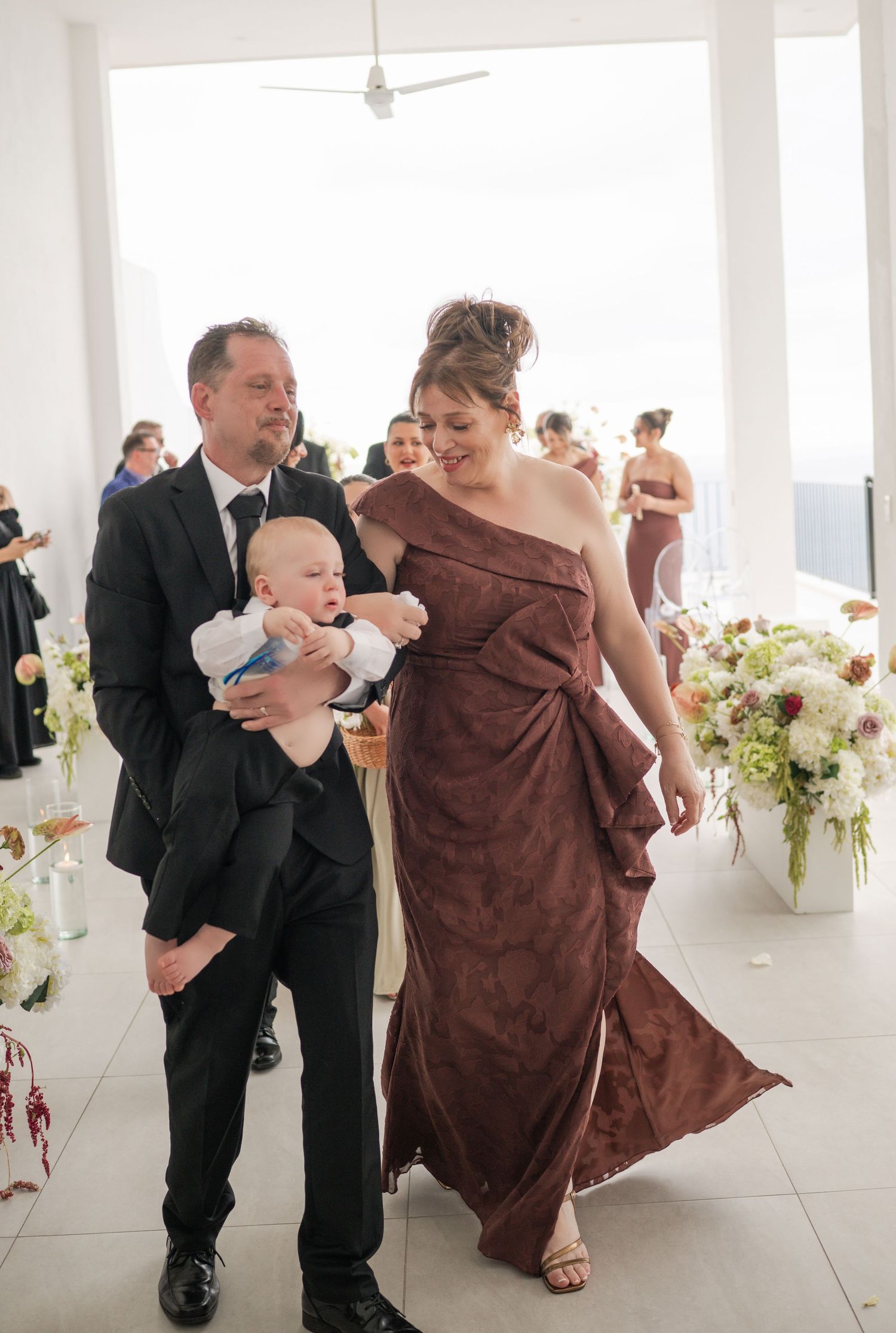 Man in black suit holds baby while woman in brown one-shoulder gown walks beside him at wedding ceremony.