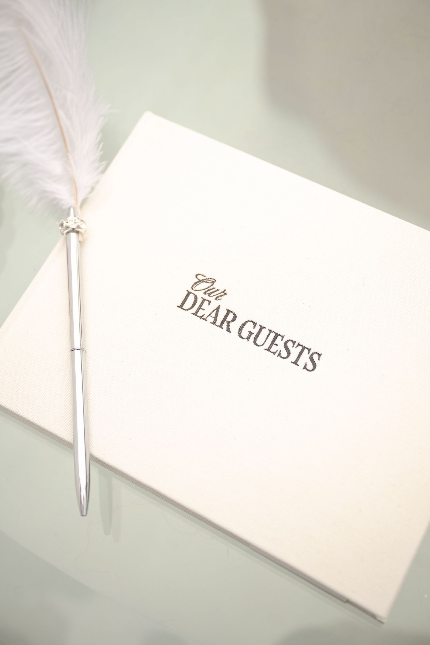 Elegant white wedding guest book with feather pen on glass table, reading Our Dear Guests in gold script.