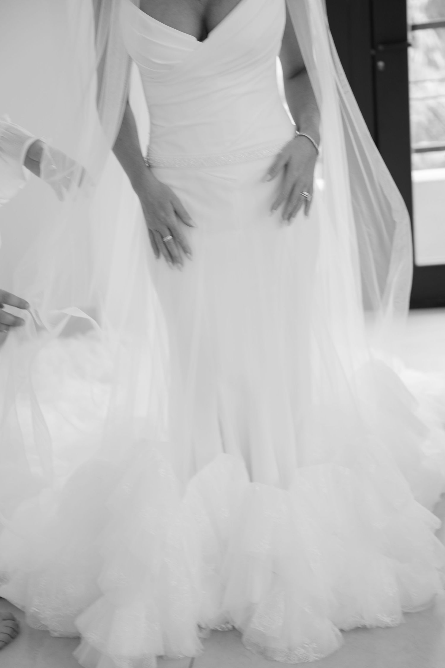 Bride in elegant off-shoulder ruched tulle wedding gown with ruffled hem and flowing veil, black and white photo.