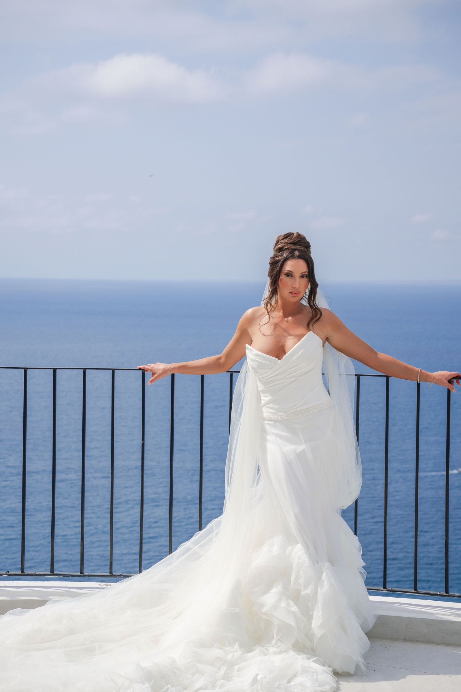 Bride in a flowing white wedding gown poses on a seaside balcony with stunning ocean views.
