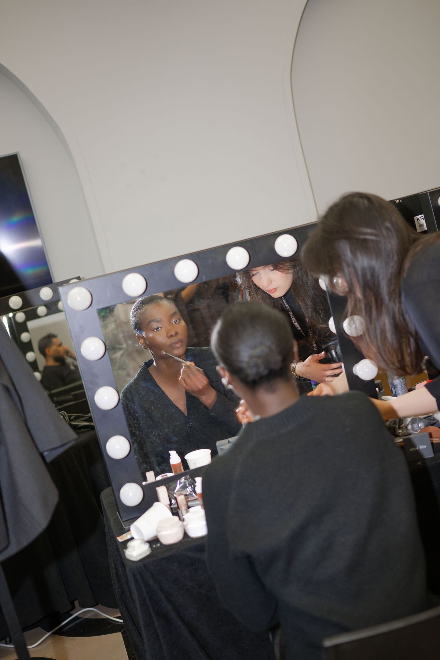 Makeup artist applying cosmetics to a model at a Hollywood vanity mirror with lights in a backstage dressing room.