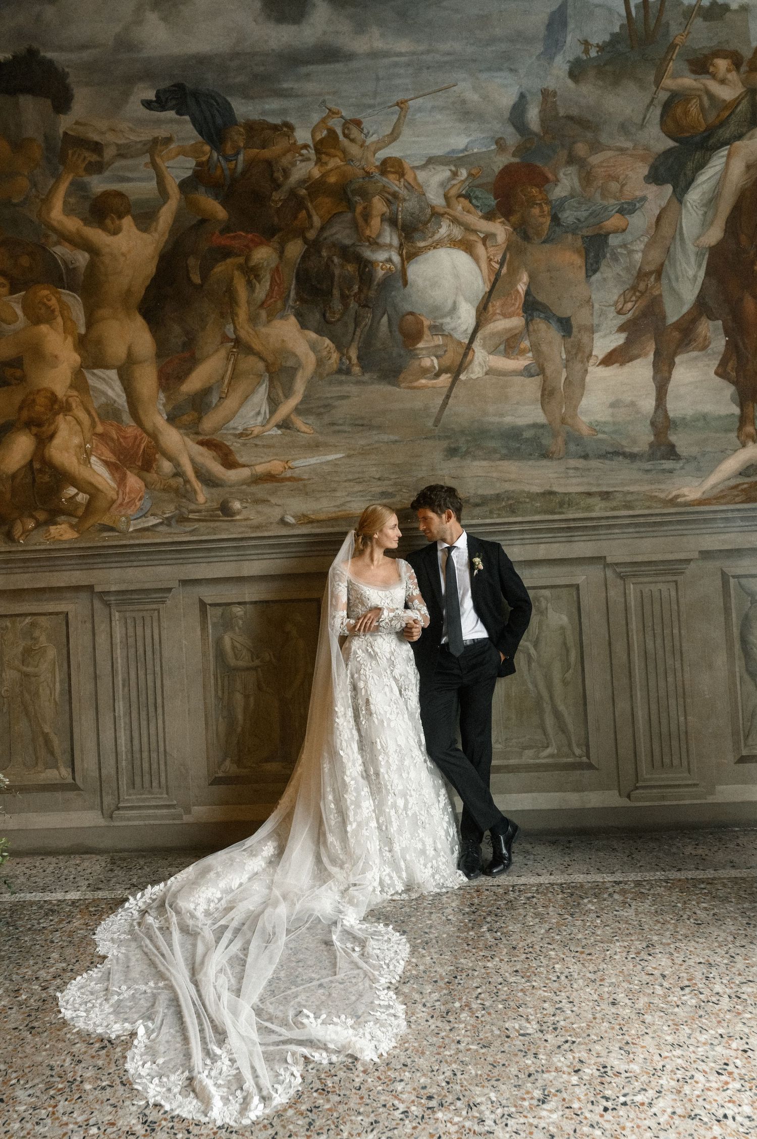 Bride in lace gown with veil and groom in tuxedo pose before a classical fresco mural in an elegant Italian villa.