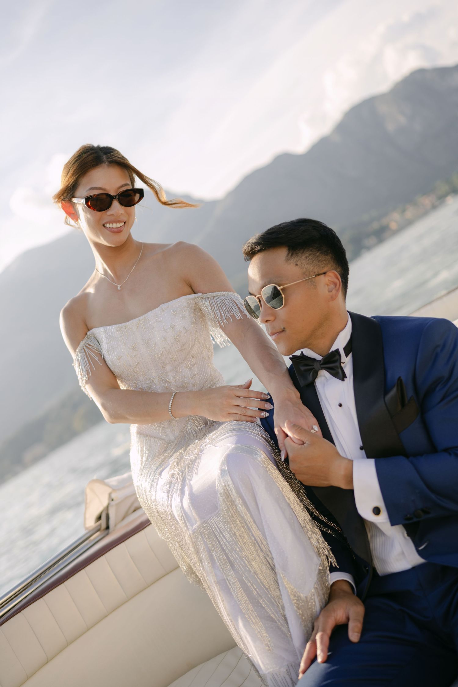 Couple in wedding attire on a boat with mountain lake backdrop, bride in white gown and groom in navy tuxedo.