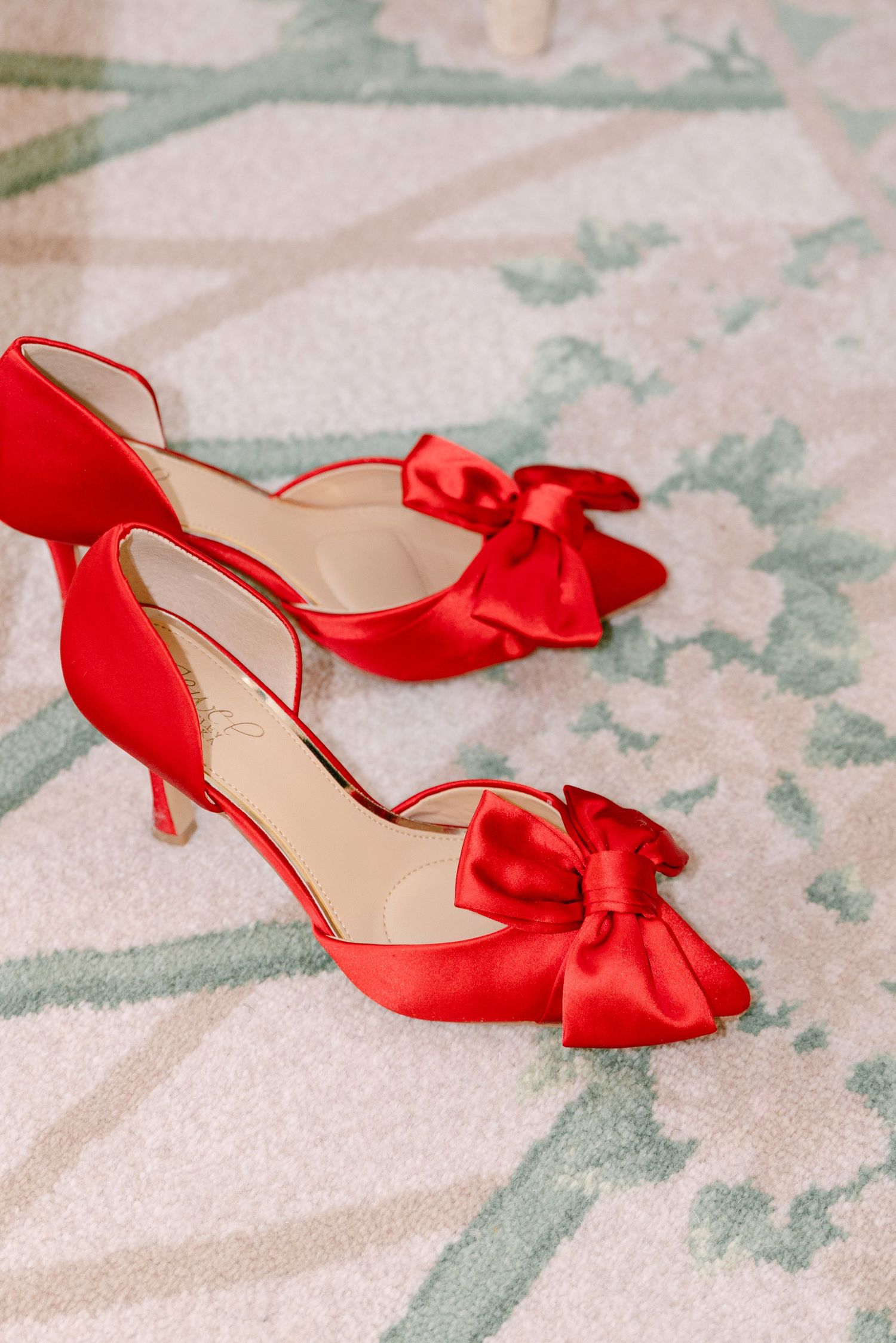 Red satin kitten heel wedding shoes with large bow detail on floral patterned carpet.