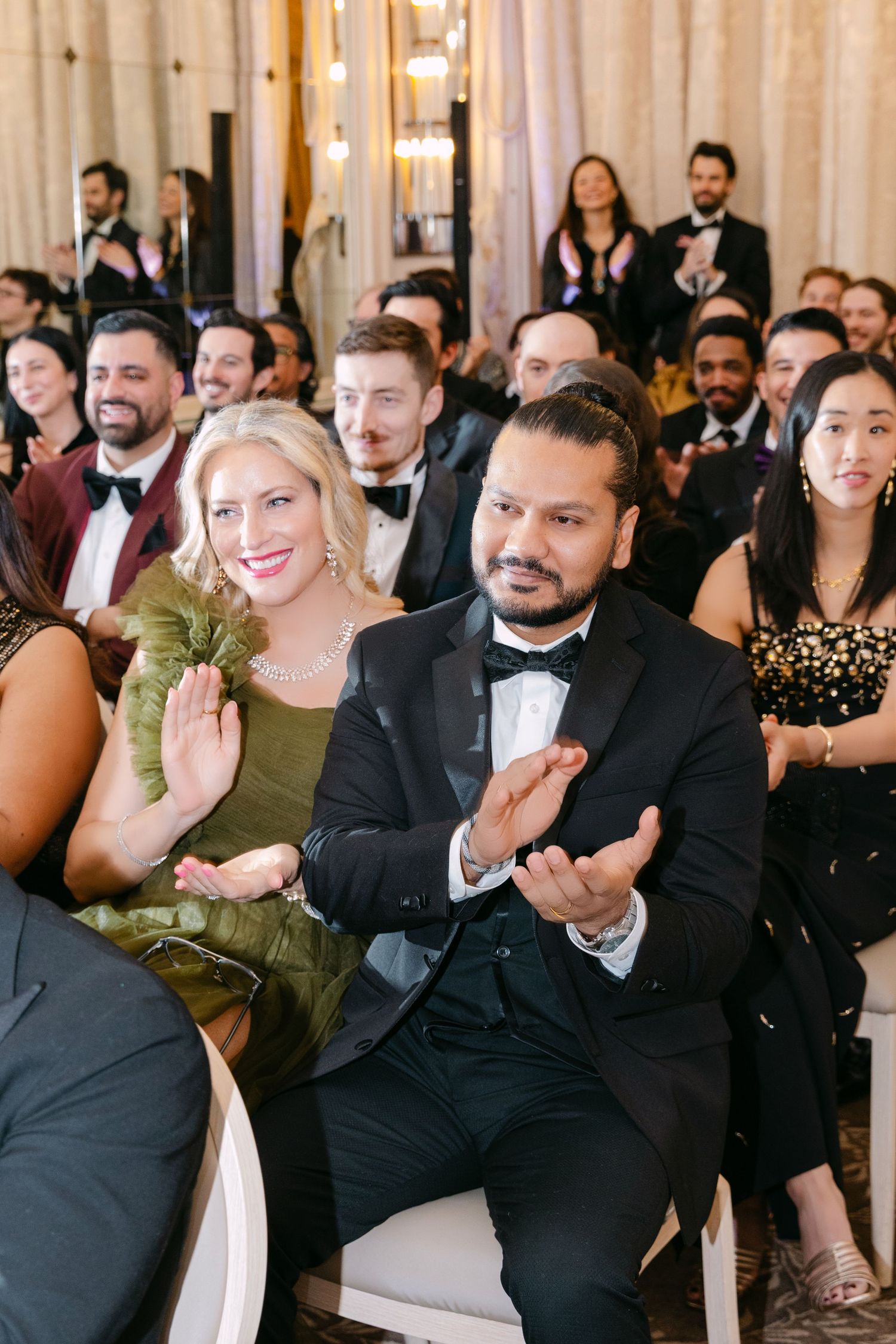 Wedding guests in formal attire applauding during ceremony in a grand ballroom with chandeliers and mirrors.