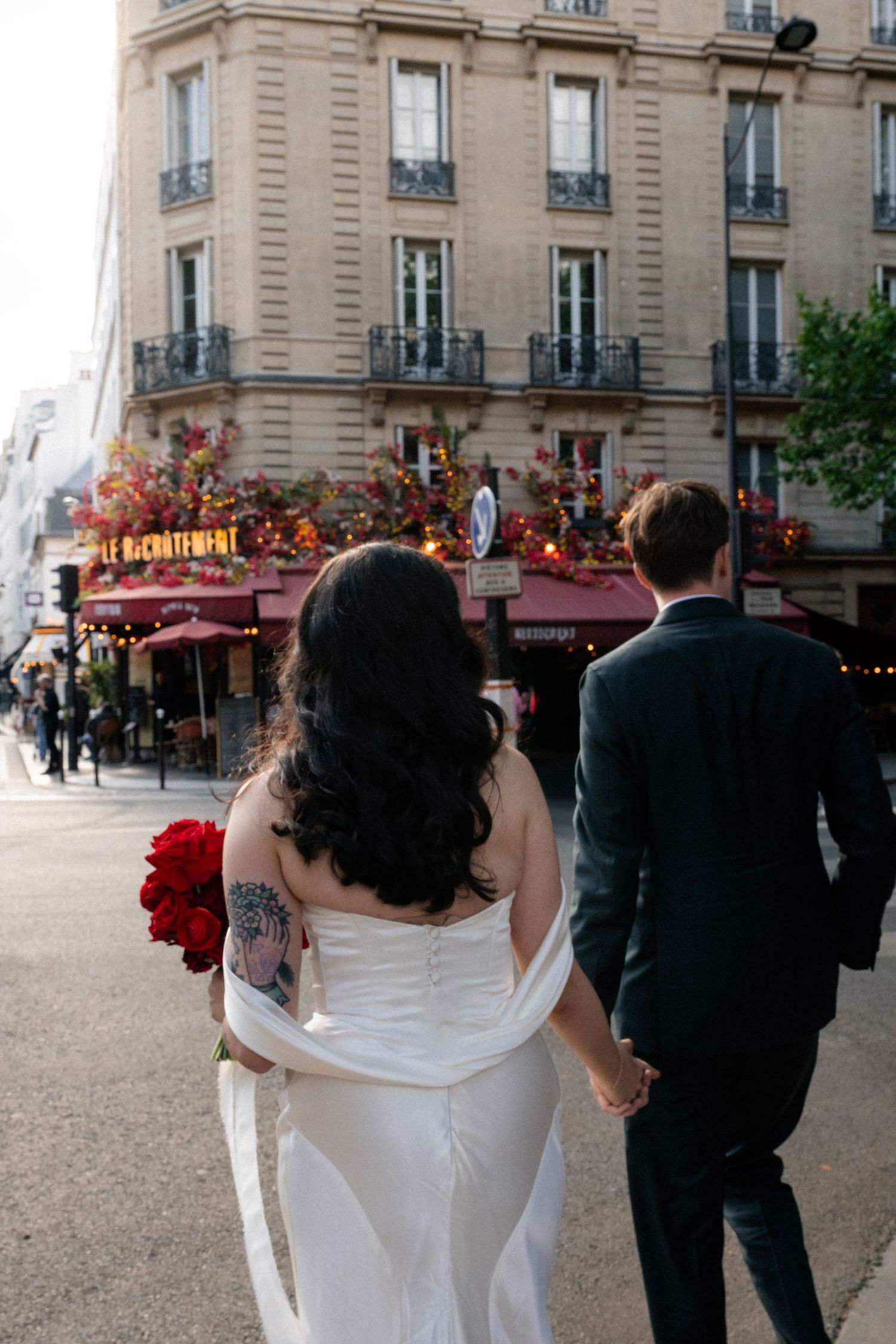 Bride with red bouquet and groom walking hand in hand through vibrant Paris street cafe district.