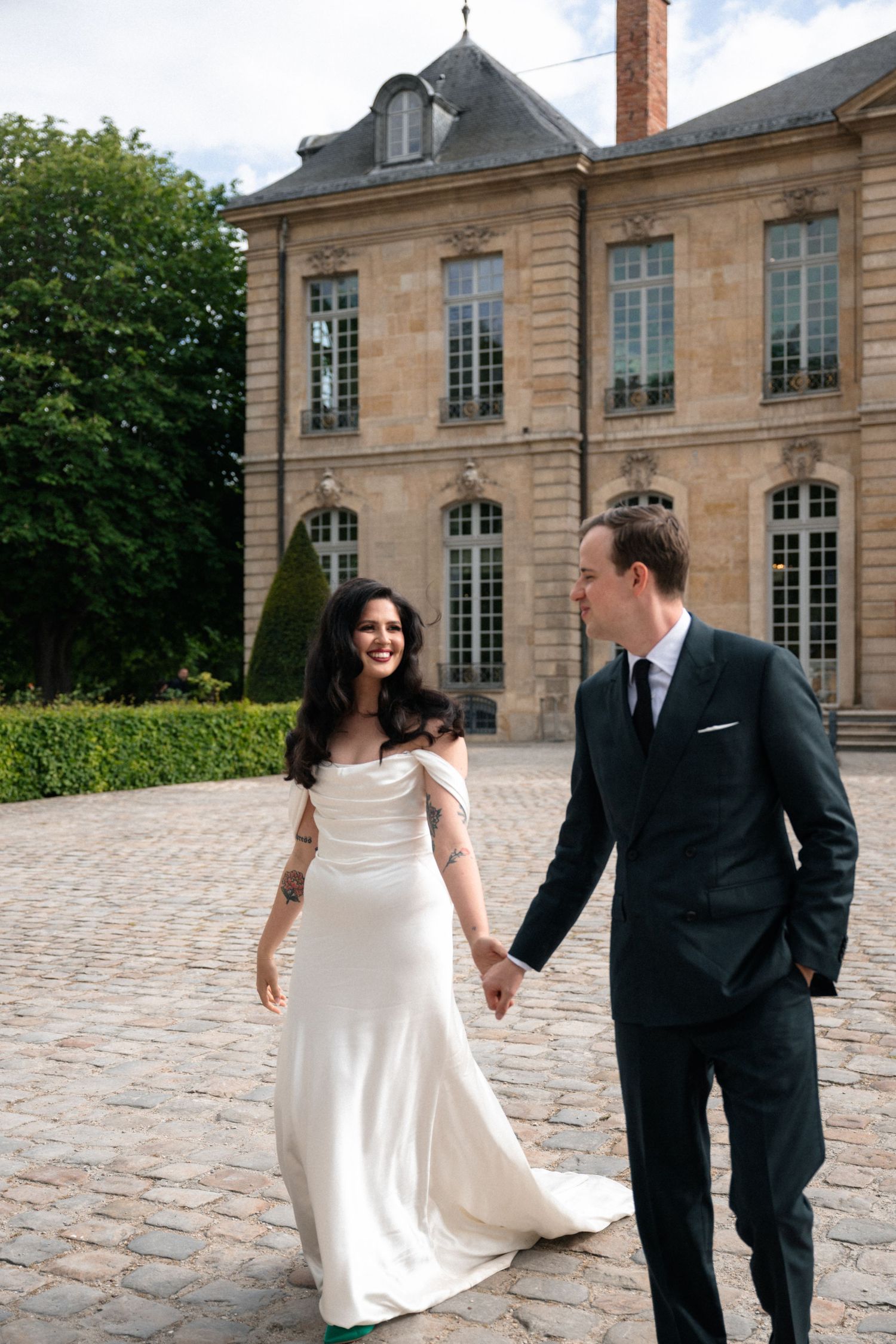 Bride and groom holding hands walking on cobblestone courtyard in front of elegant French chateau on wedding day.