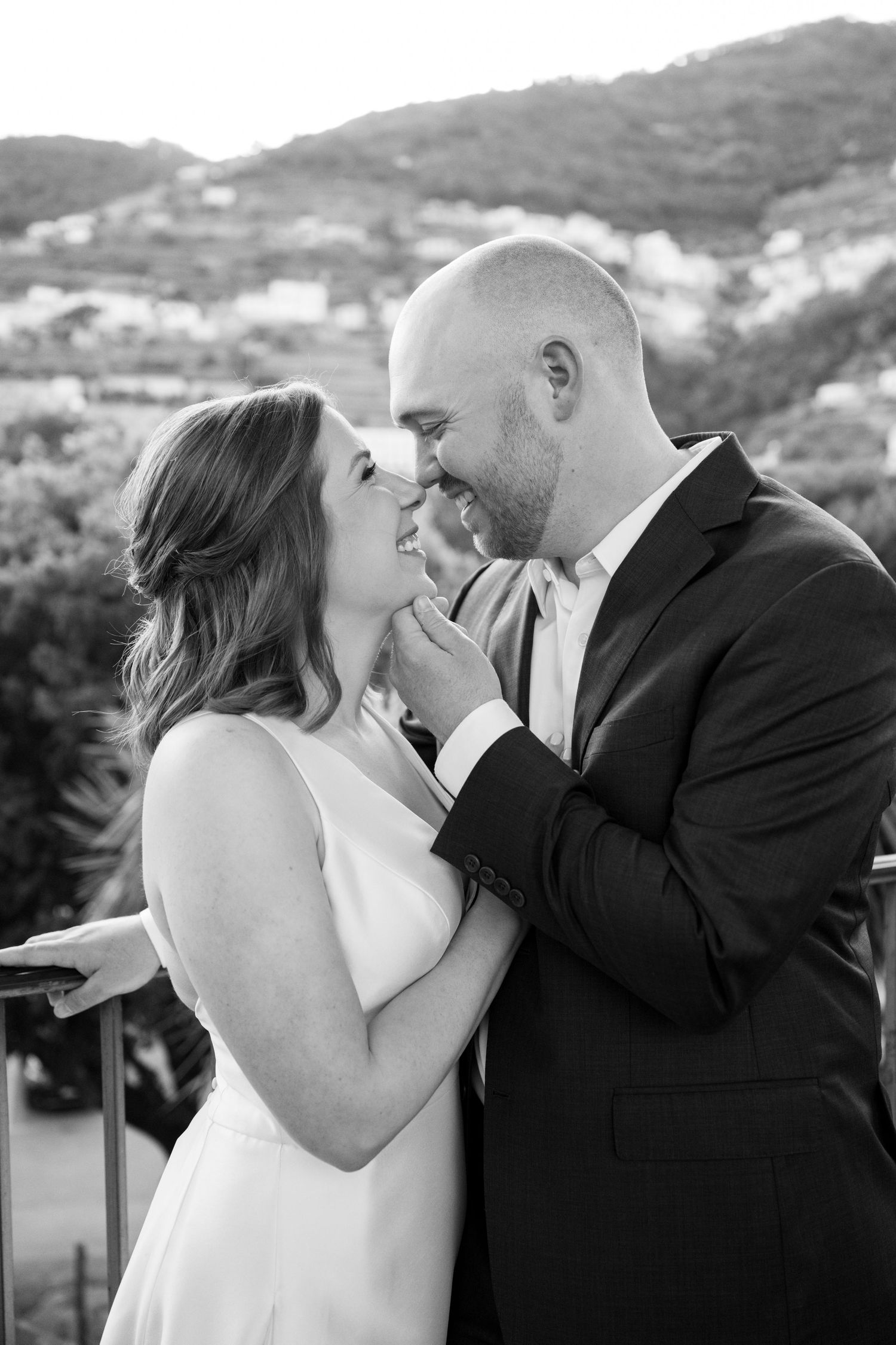 Black and white photo of a smiling couple sharing a romantic moment on a balcony with mountain views.