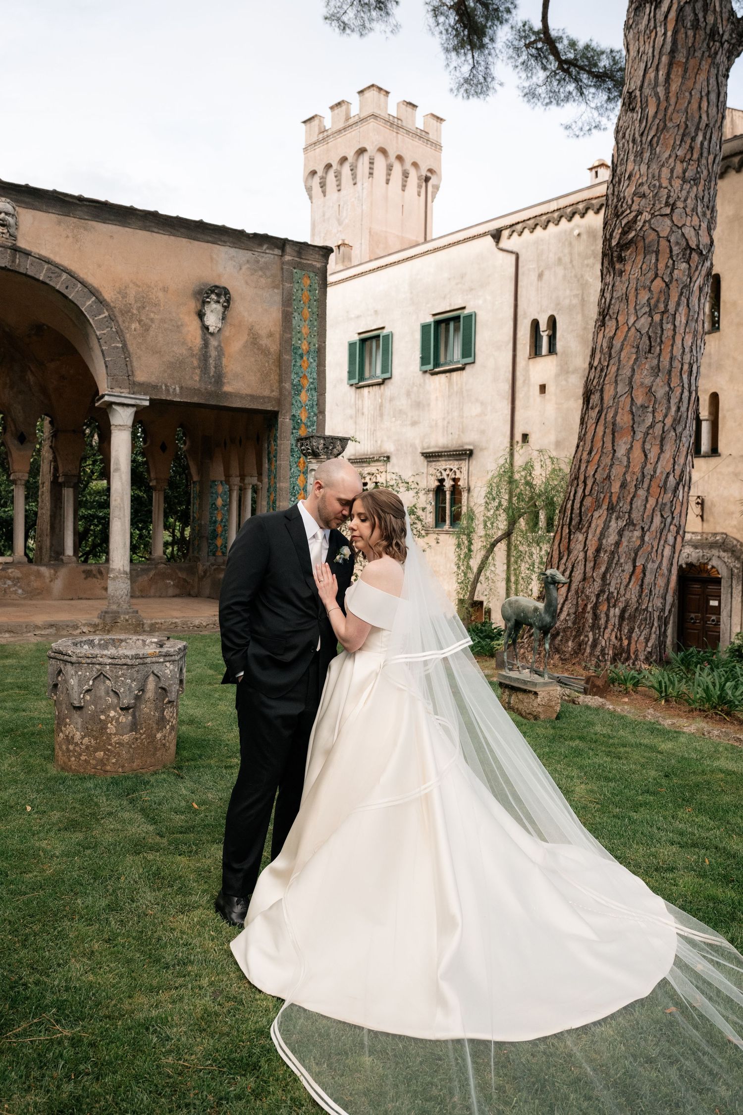 Bride and groom embrace in the garden of a historic Italian castle with flowing veil and lush greenery.