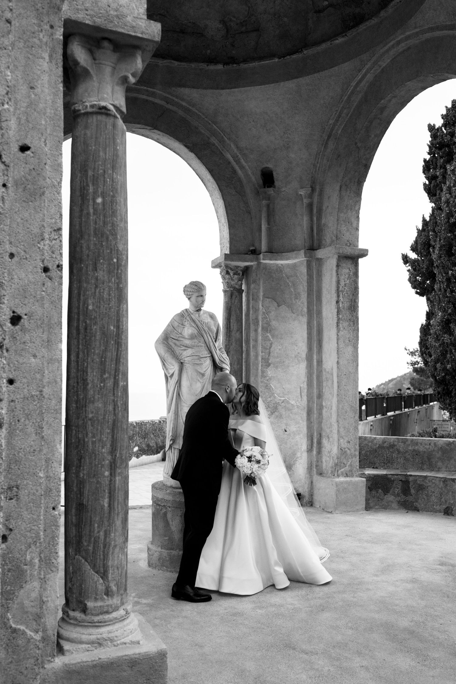 Black and white photo of bride and groom sharing a kiss under ancient stone arches with a classical statue nearby.