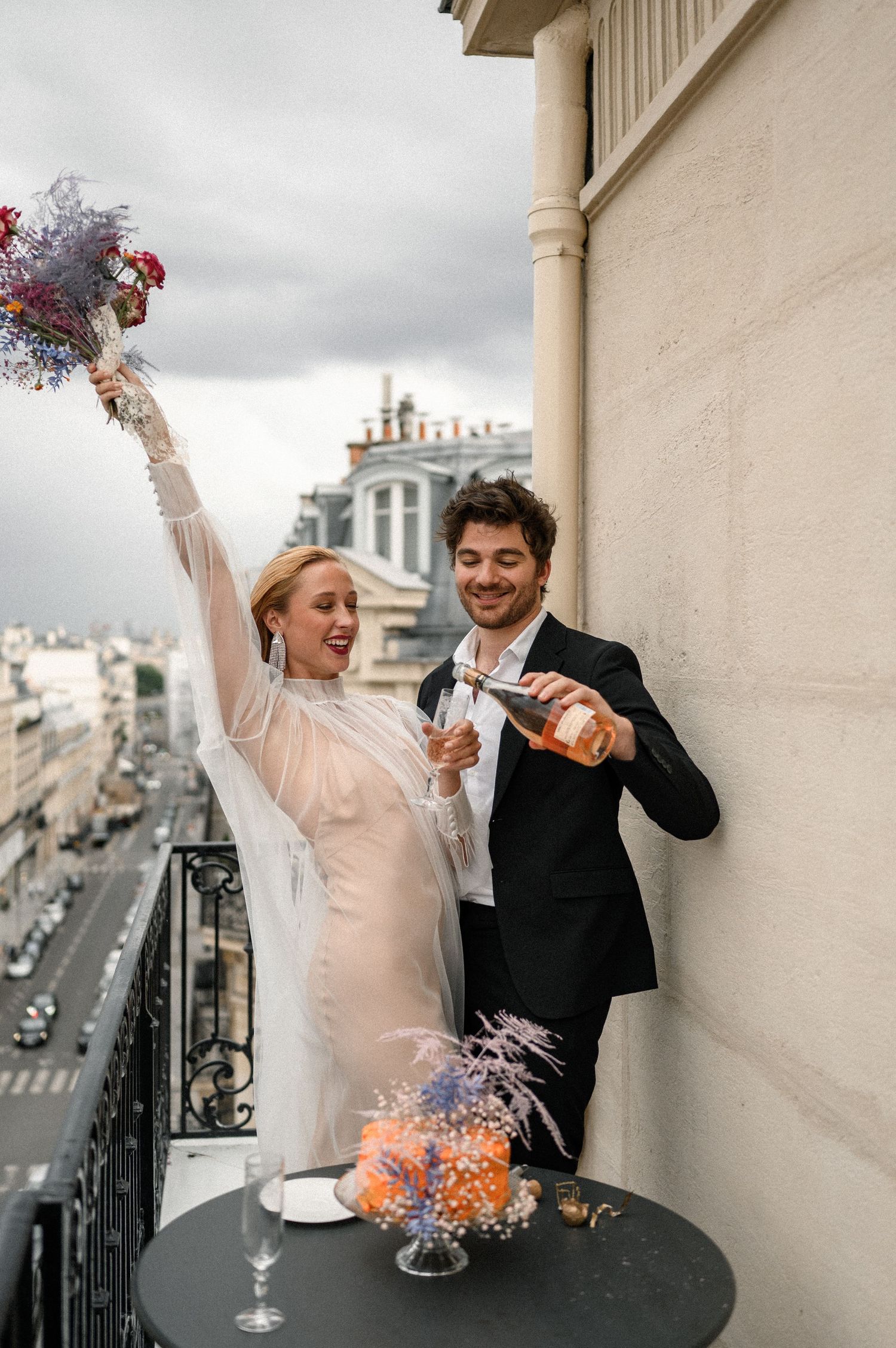 Joyful bride raising bouquet while groom pours champagne on elegant Paris hotel balcony with city skyline.