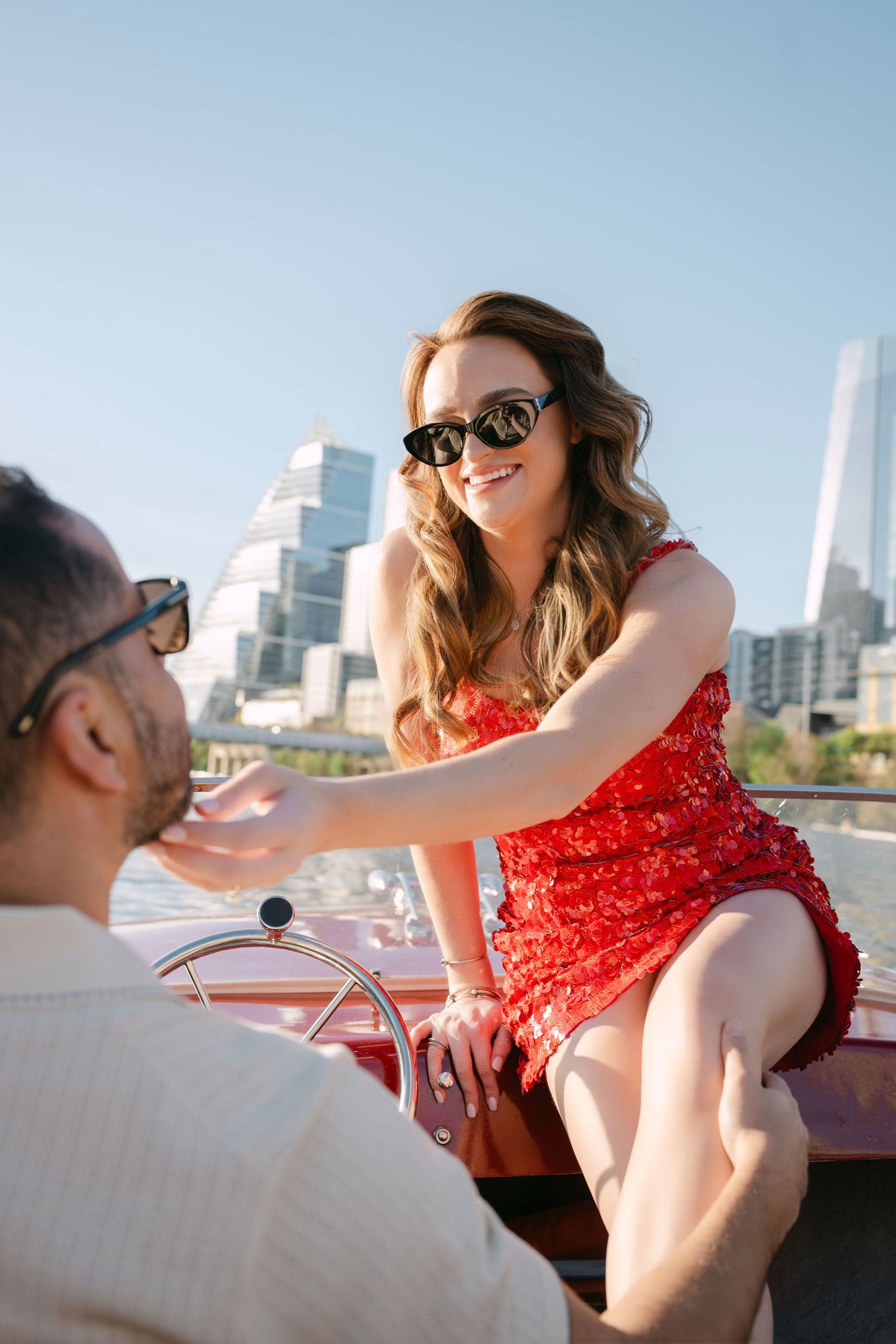 Smiling woman in red sequin dress and sunglasses sitting on a classic red boat with Austin Texas skyline behind her.