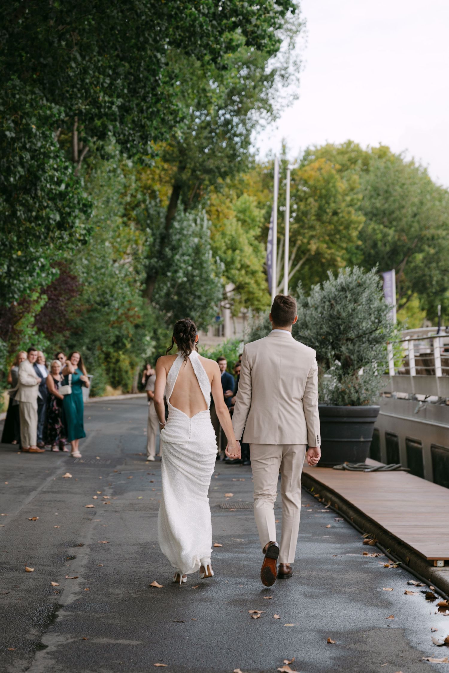 Bride and groom walk hand in hand down a tree-lined path while wedding guests look on.