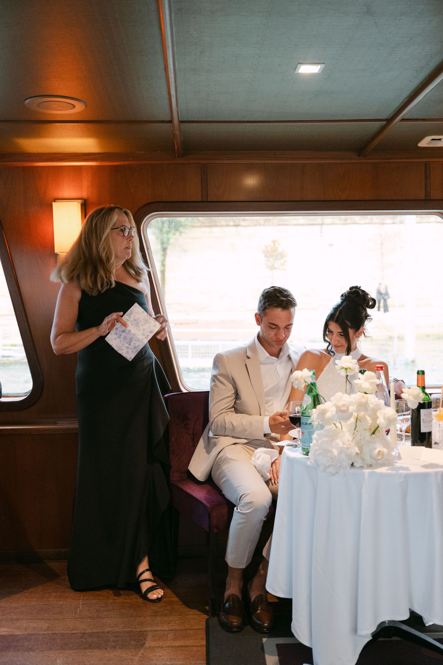 A woman gives a wedding speech while the bride and groom sit at their decorated table on a boat.
