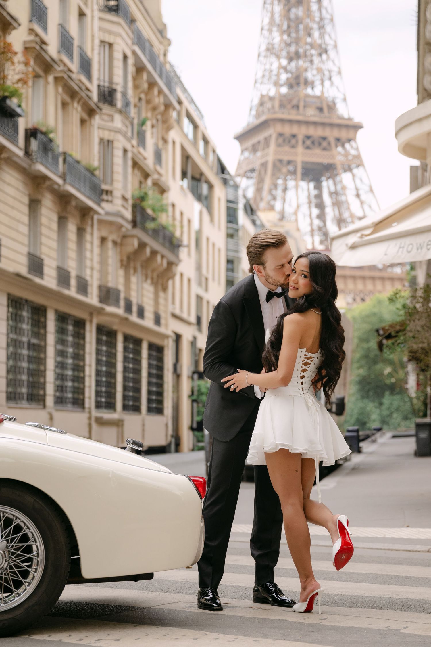 Elegant couple embracing near vintage white car with Eiffel Tower backdrop in Paris, woman in white mini dress with red heels.